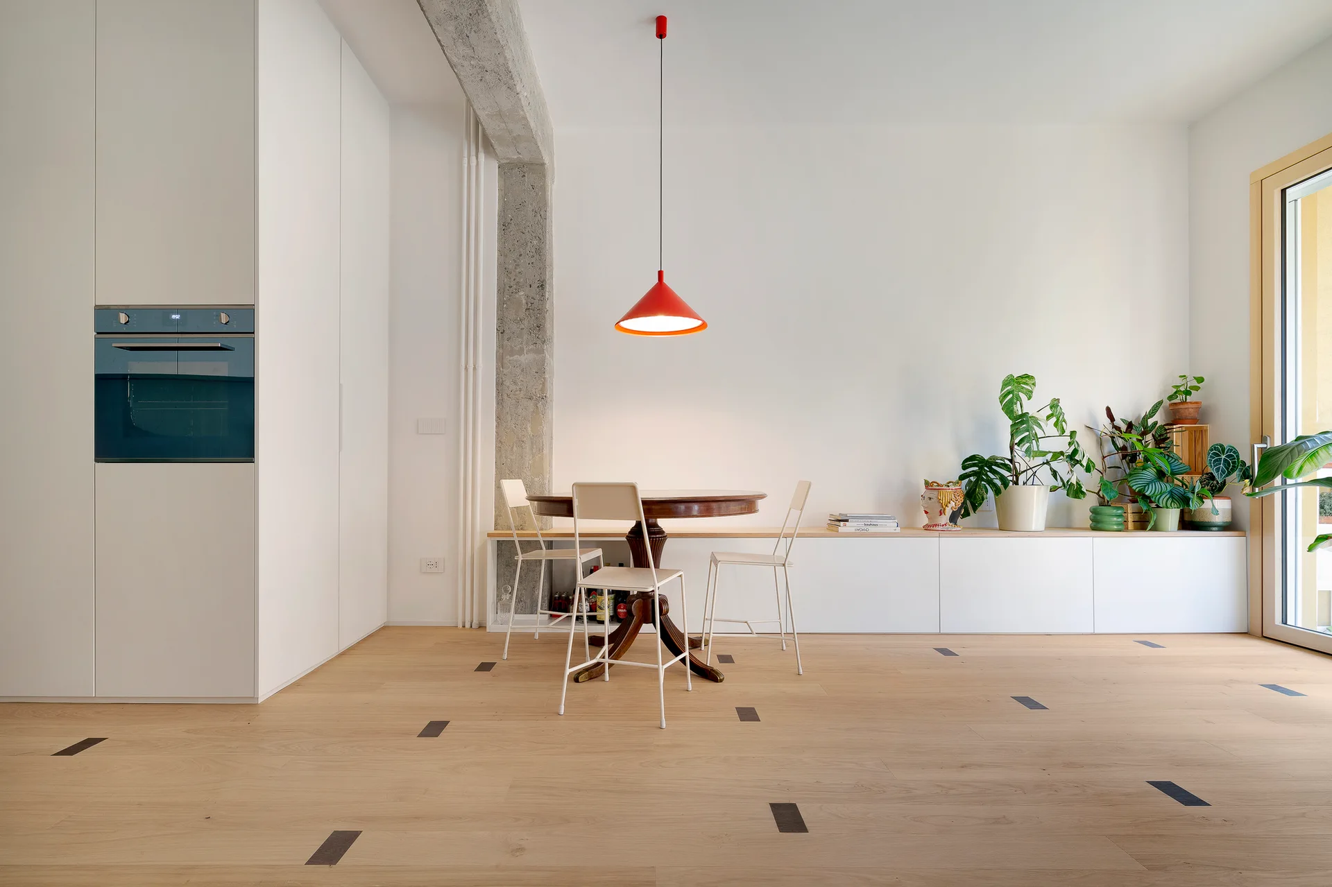 A minimalist dining room featuring a round wooden table, white metal chairs, a red pendant light, and an exposed concrete column.