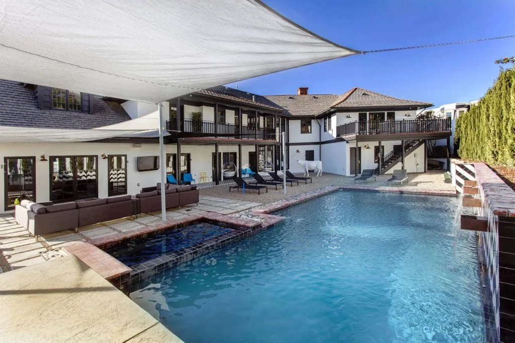 A large blue swimming pool with a waterfall is surrounded by a gravel patio with lounge chairs and a covered sectional sofa under a white shade sail.