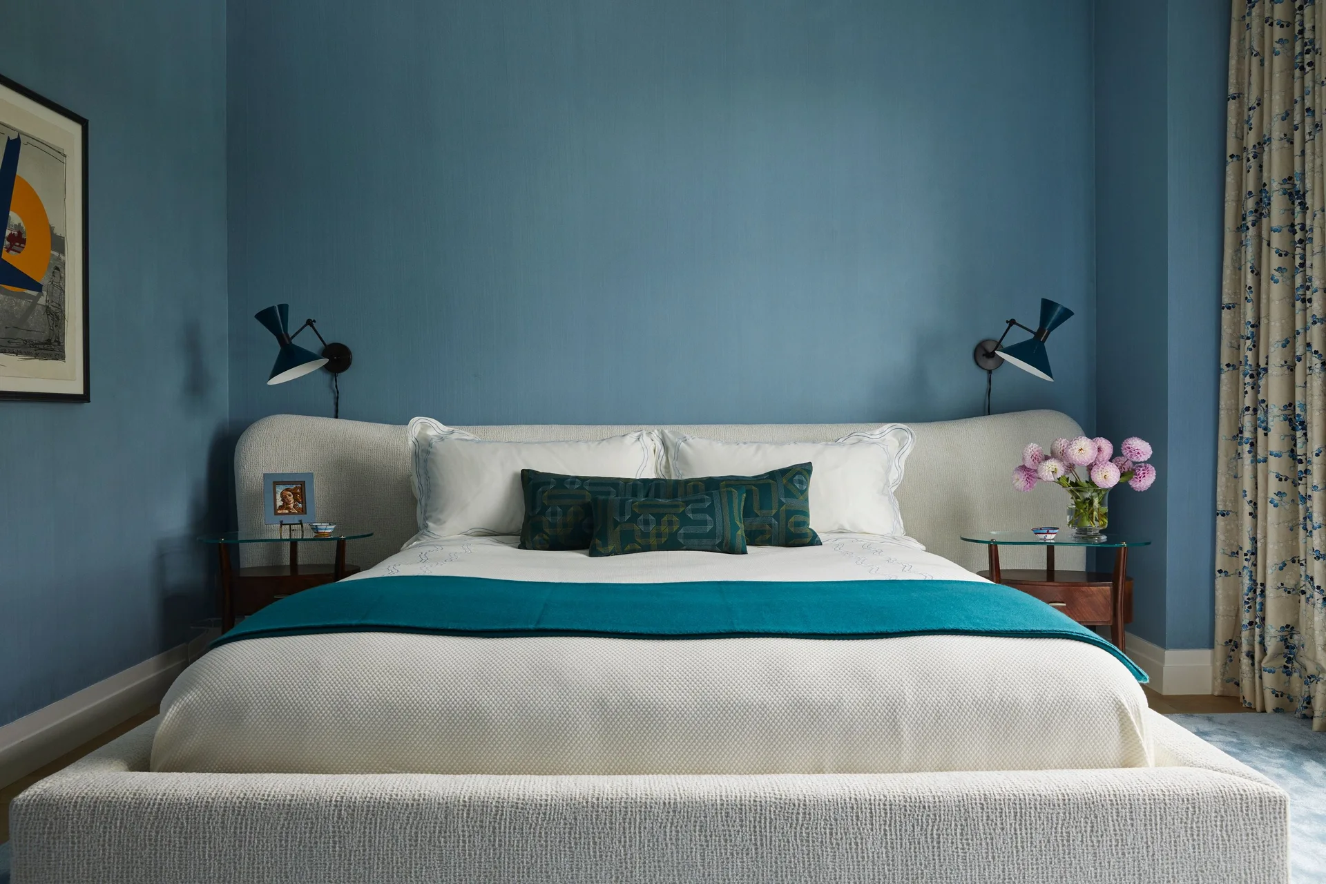 A symmetrical bedroom with blue textured wallpaper, a curved cream upholstered bed, glass-top nightstands, and matching blue wall sconces.