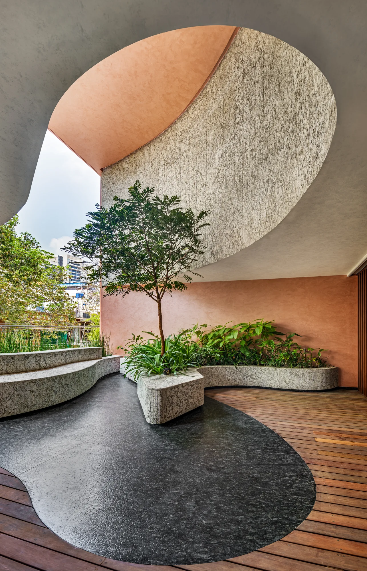 An outdoor terrace features curved stone built-in benches, a central tree in a planter, a wood plank floor with a dark stone inlay, and terracotta walls.