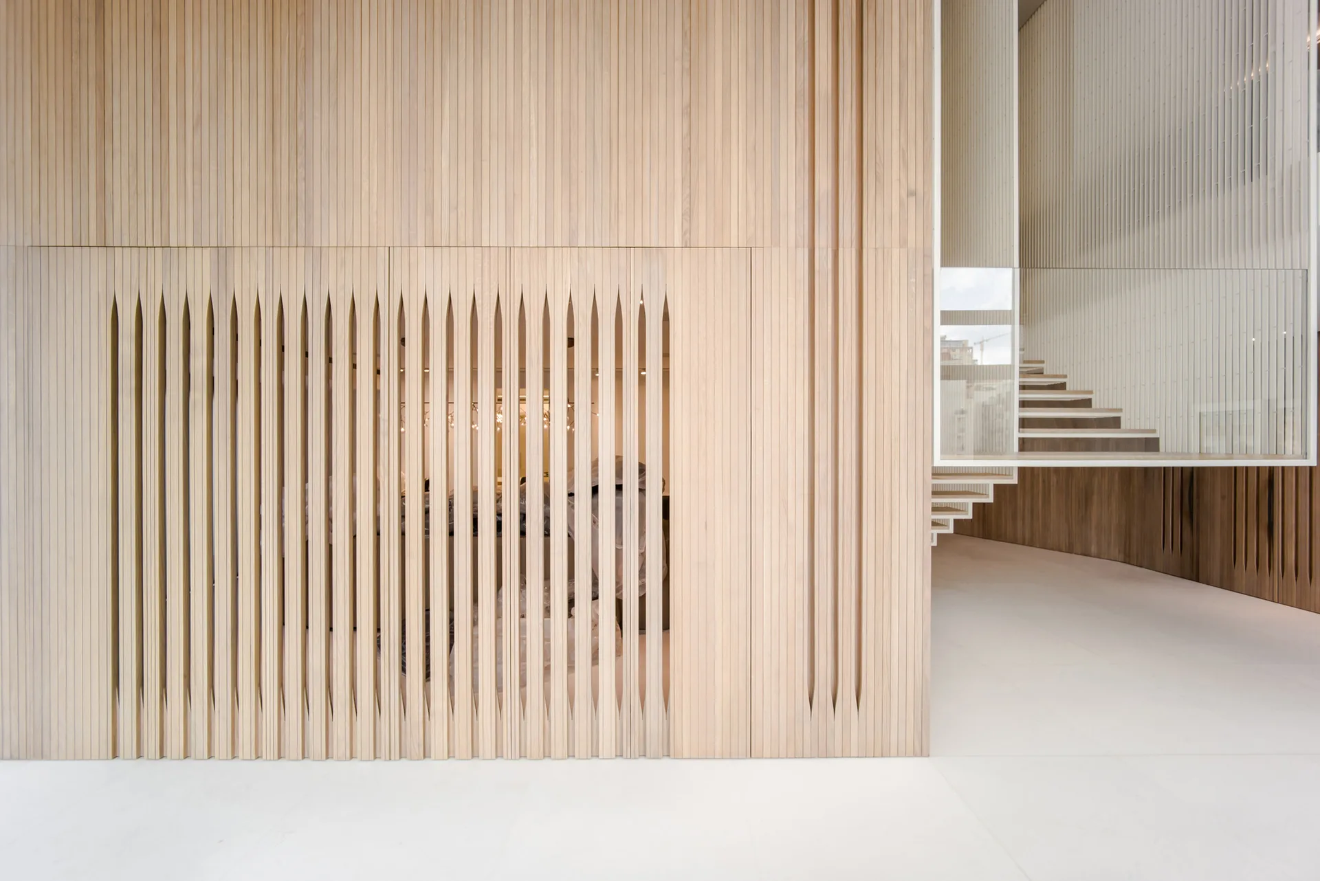 A minimalist interior featuring a floor-to-ceiling slatted light wood wall with decorative vertical cutouts next to a floating staircase with glass railings.