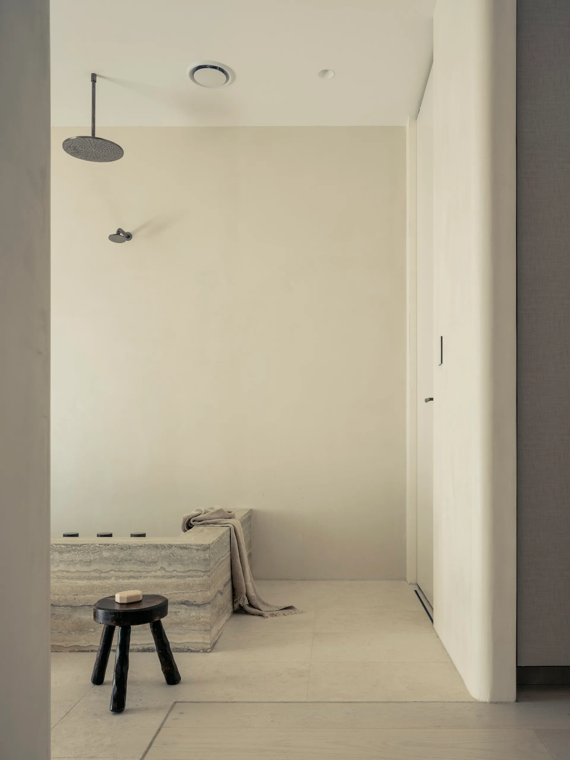 A minimalist bathroom features a rectangular veined stone bathtub, a small dark wooden stool, and smooth beige plaster walls.