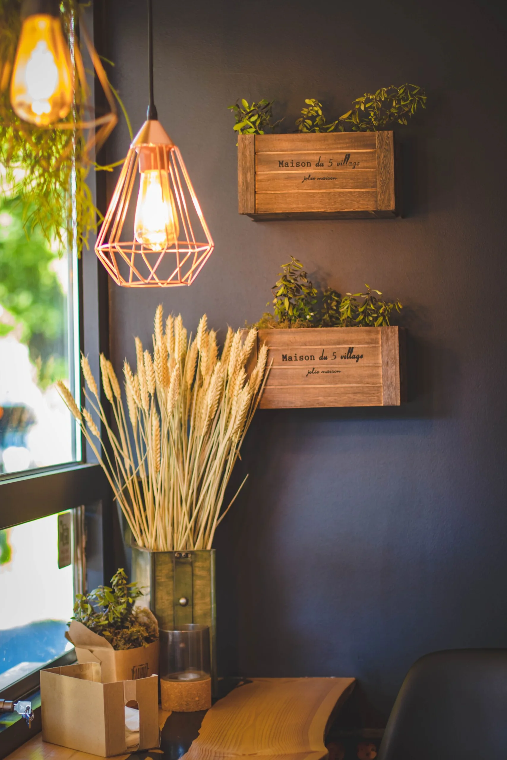 A dark grey wall decorated with wall-mounted rustic wood planters, a tall green vase of dried wheat, and a glowing copper geometric pendant light.