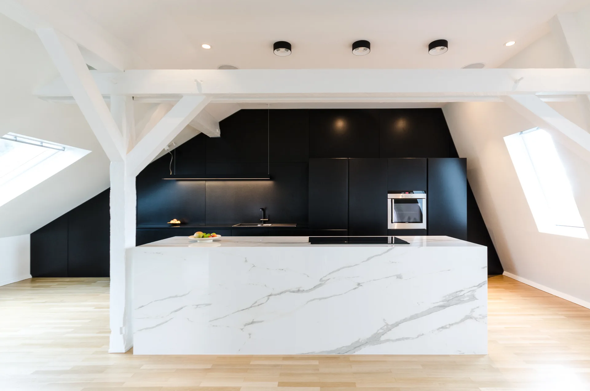 A modern attic kitchen featuring a large white marble island, matte black cabinetry beneath a sloped ceiling, and white painted beams.