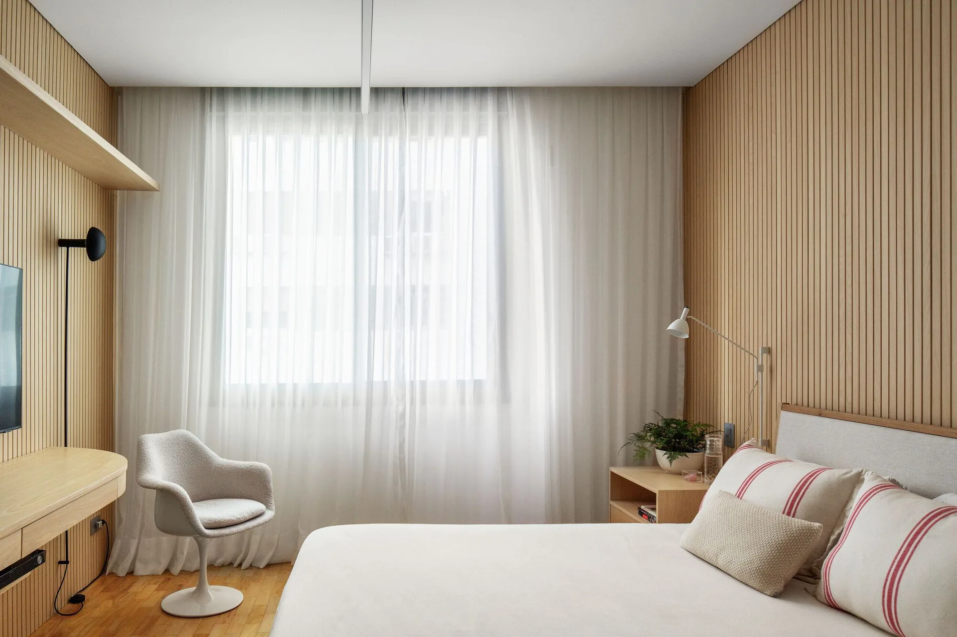 Modern bedroom featuring vertical wood slat wall paneling, a floating wooden desk, a light gray swivel chair, and sheer white curtains.