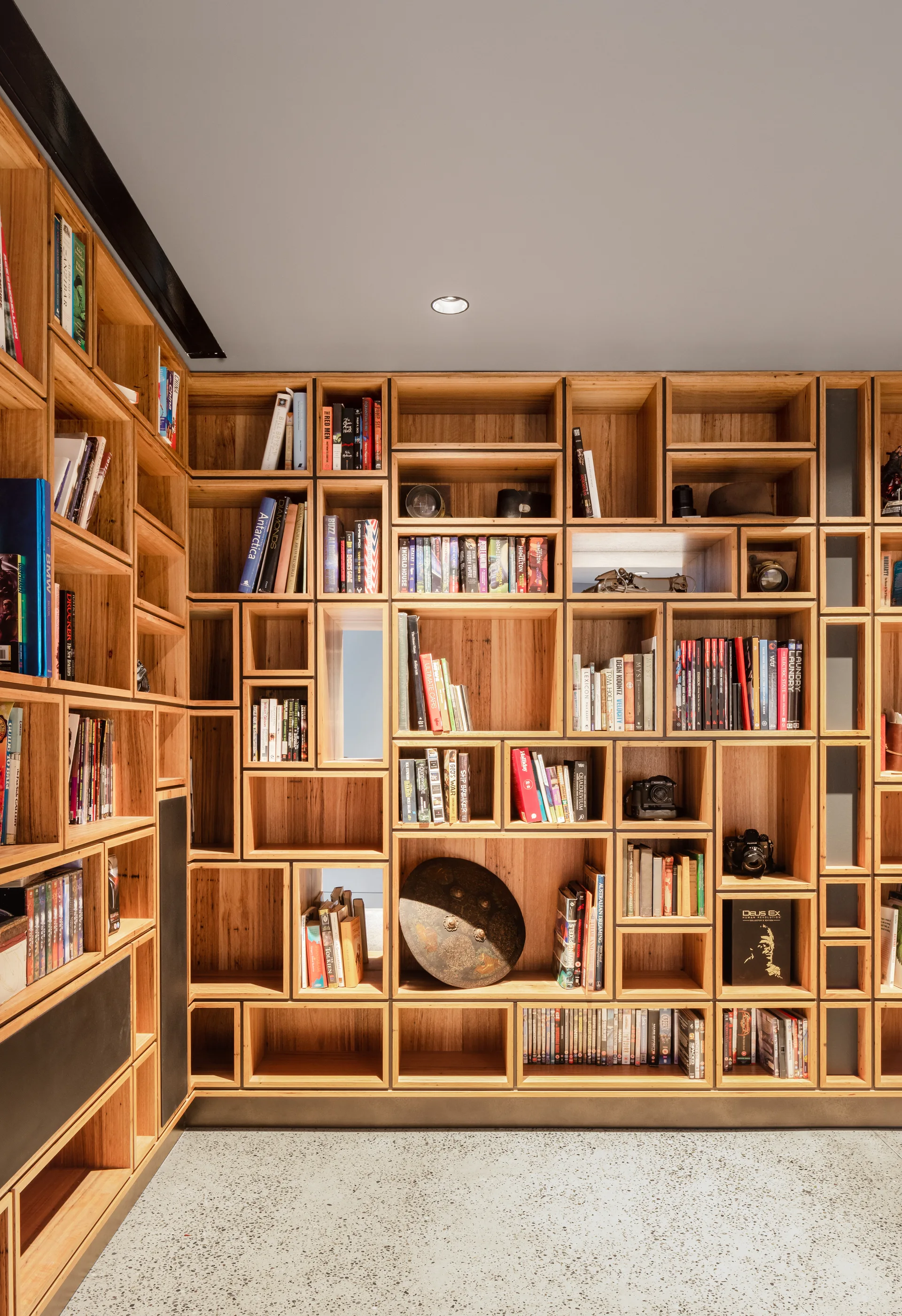 A custom floor-to-ceiling wooden bookshelf with irregular cubby storage stands on a speckled grey terrazzo floor.