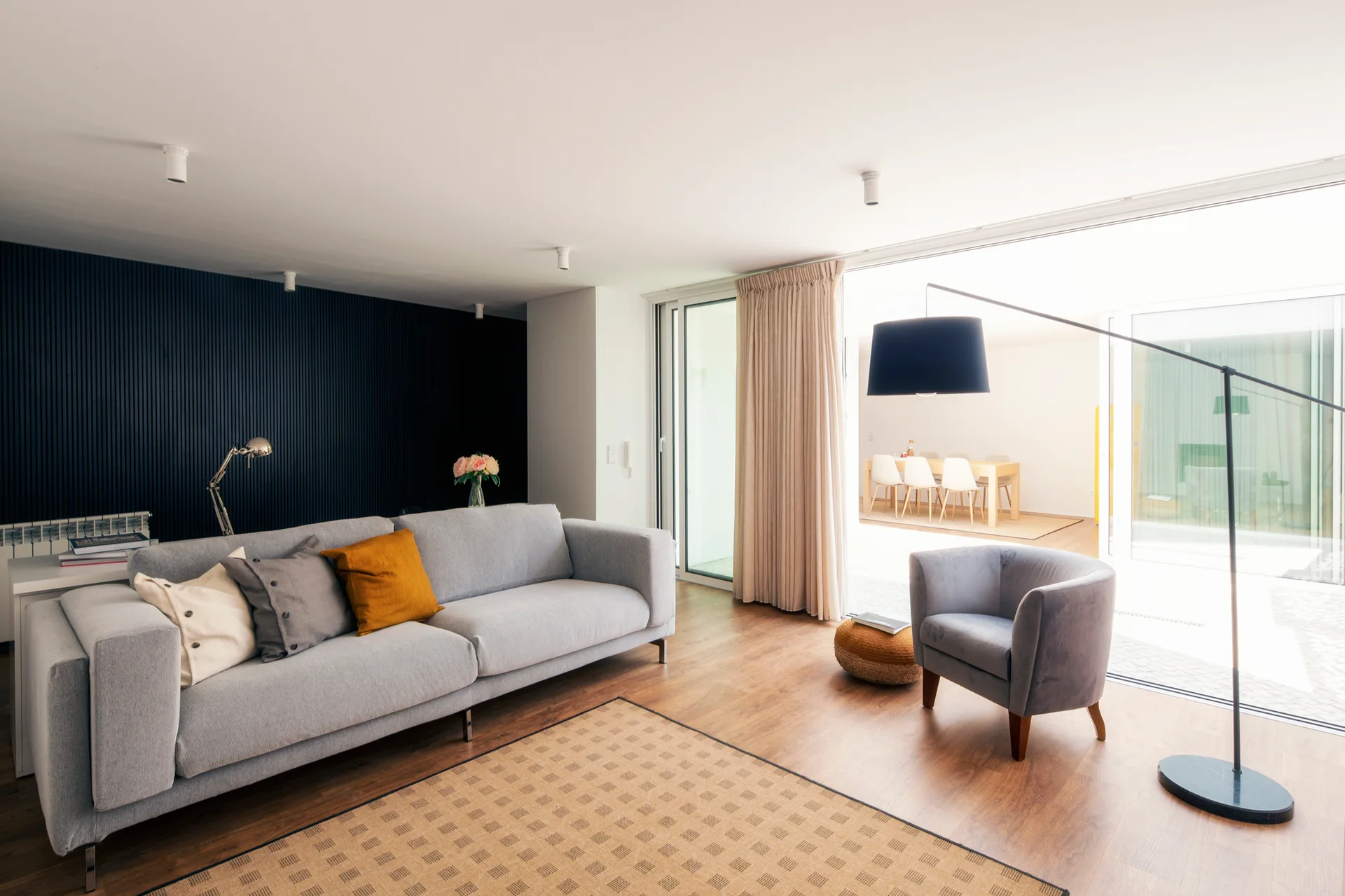 A contemporary living room featuring a grey sofa set against a dark blue slatted accent wall, alongside an armchair and large glass doors.