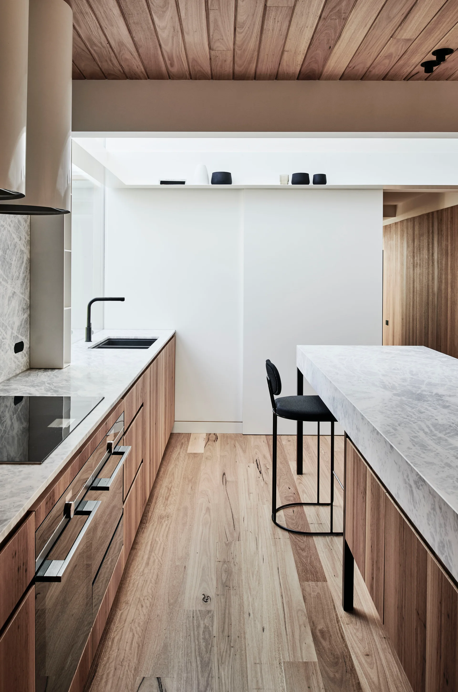 A minimalist kitchen features light wood cabinetry, a matching wood plank ceiling, marble countertops, and a single black bar stool.