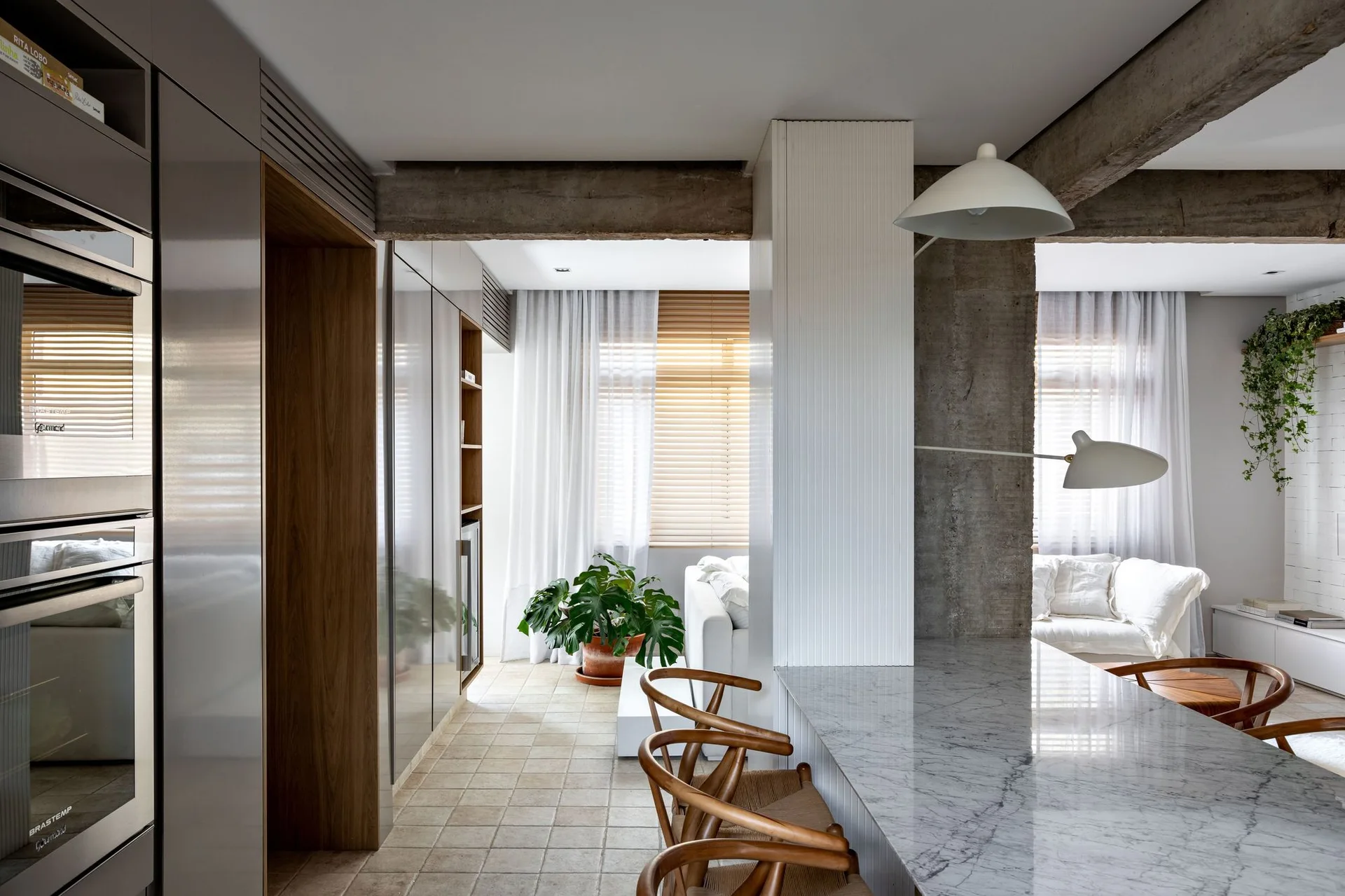An open concept kitchen features a grey marble island with wooden chairs beneath exposed concrete ceiling beams with a living space in the background.