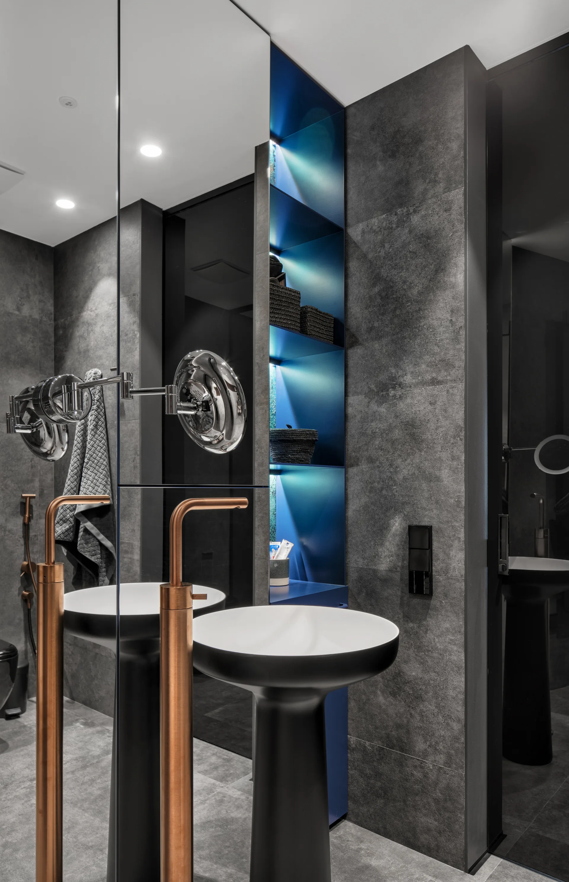 A modern bathroom features a black pedestal sink with a copper faucet, a large mirror, and a recessed vertical shelf backlit with blue light.