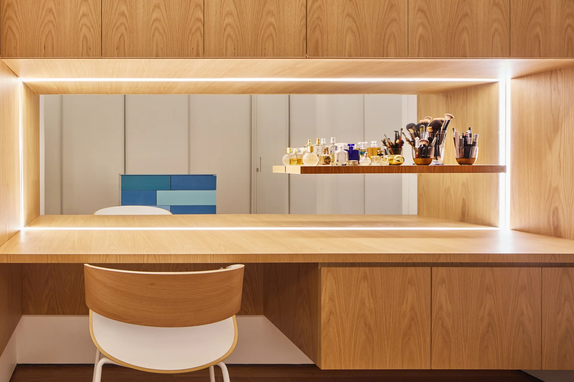 A built-in light wood vanity features a rectangular mirror bordered by LED strip lights, a floating shelf with cosmetics, and a curved wooden chair.
