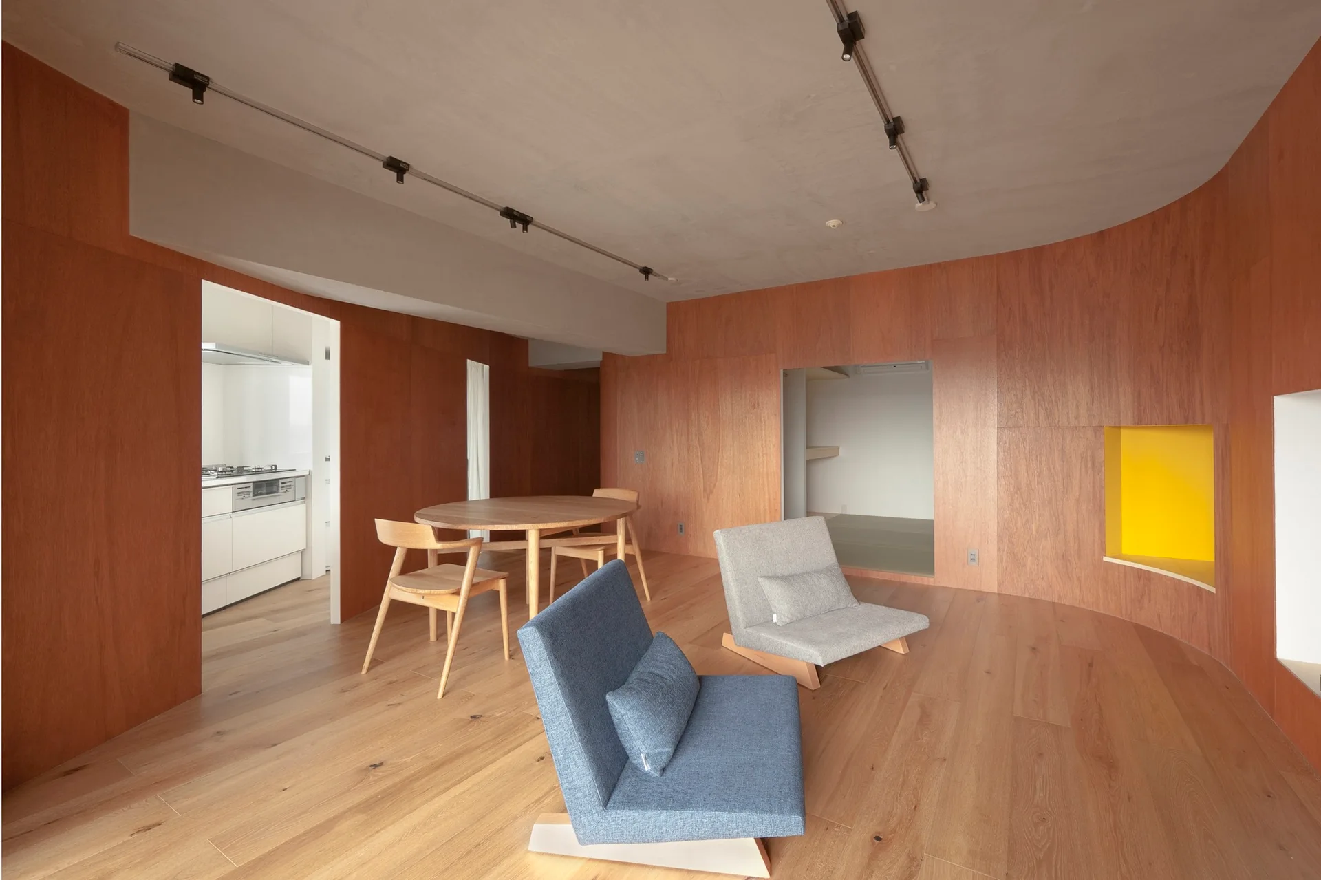 A modern living space with curved wood-paneled walls, low-profile lounge chairs, a round wooden dining set, and a recessed yellow wall niche.