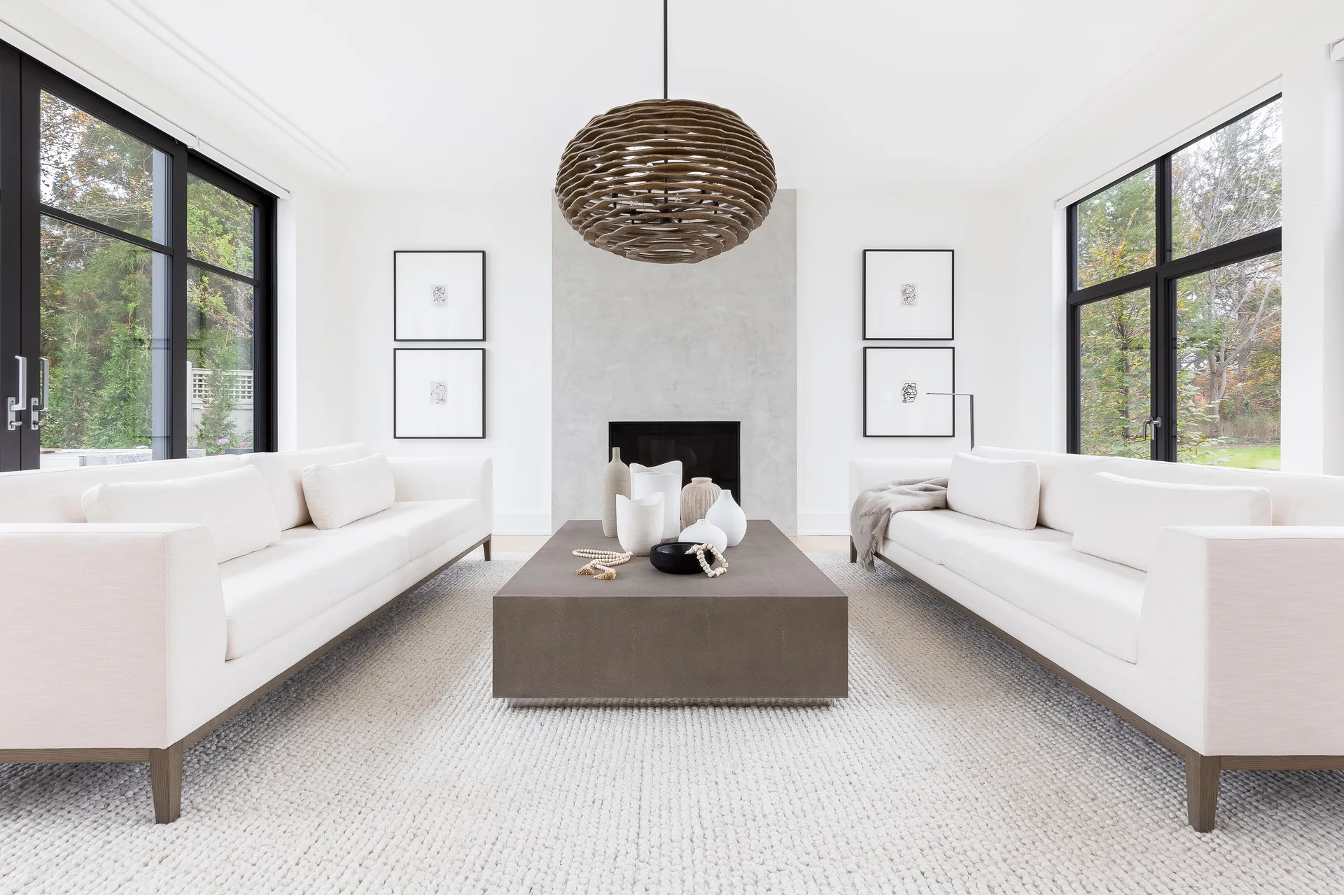 A symmetrical living room featuring two white sofas facing a low taupe coffee table on a chunky textured rug below a large wooden pendant light.