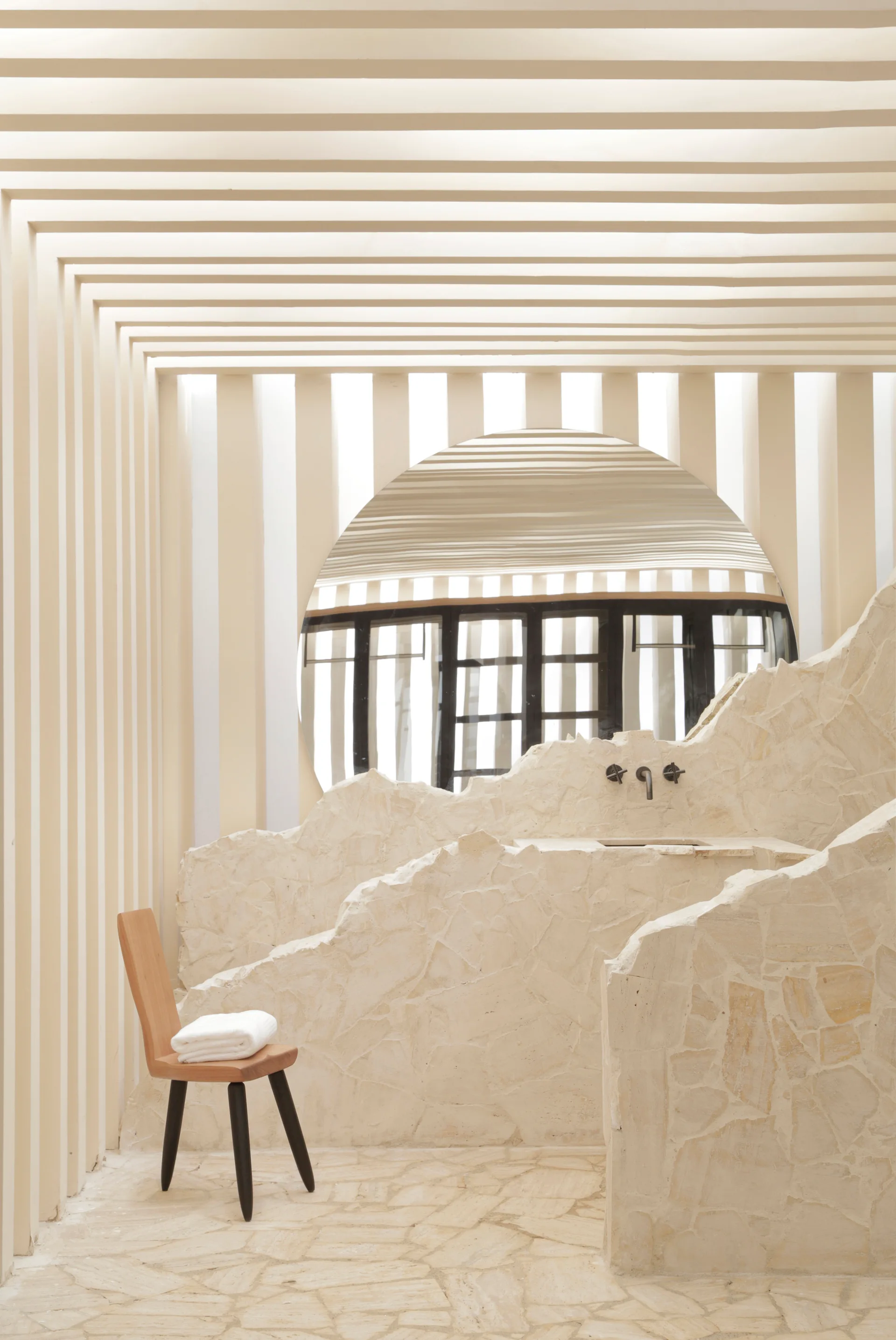 A minimalist cream bathroom featuring vertical wall slats, a large round mirror, a rough-cut stone vanity wall, and a wooden chair.