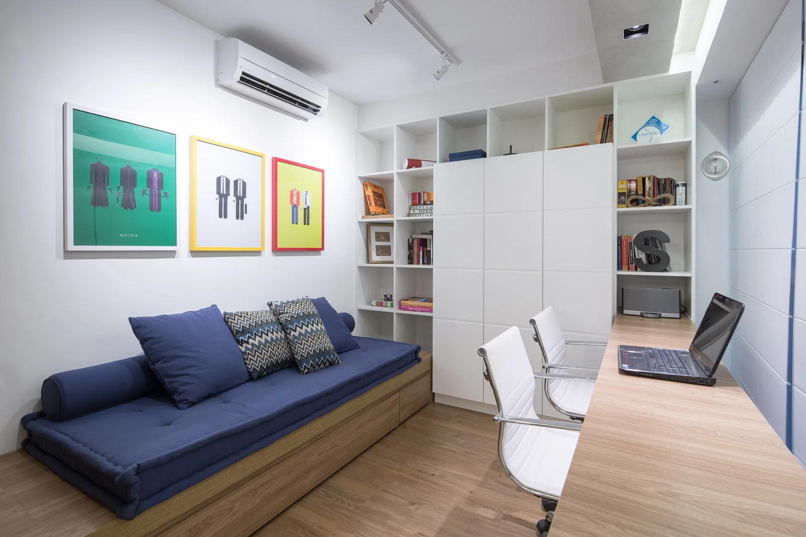 A modern home office with a long wood desk, two white swivel chairs, a blue daybed, and a wall of built-in storage shelving.