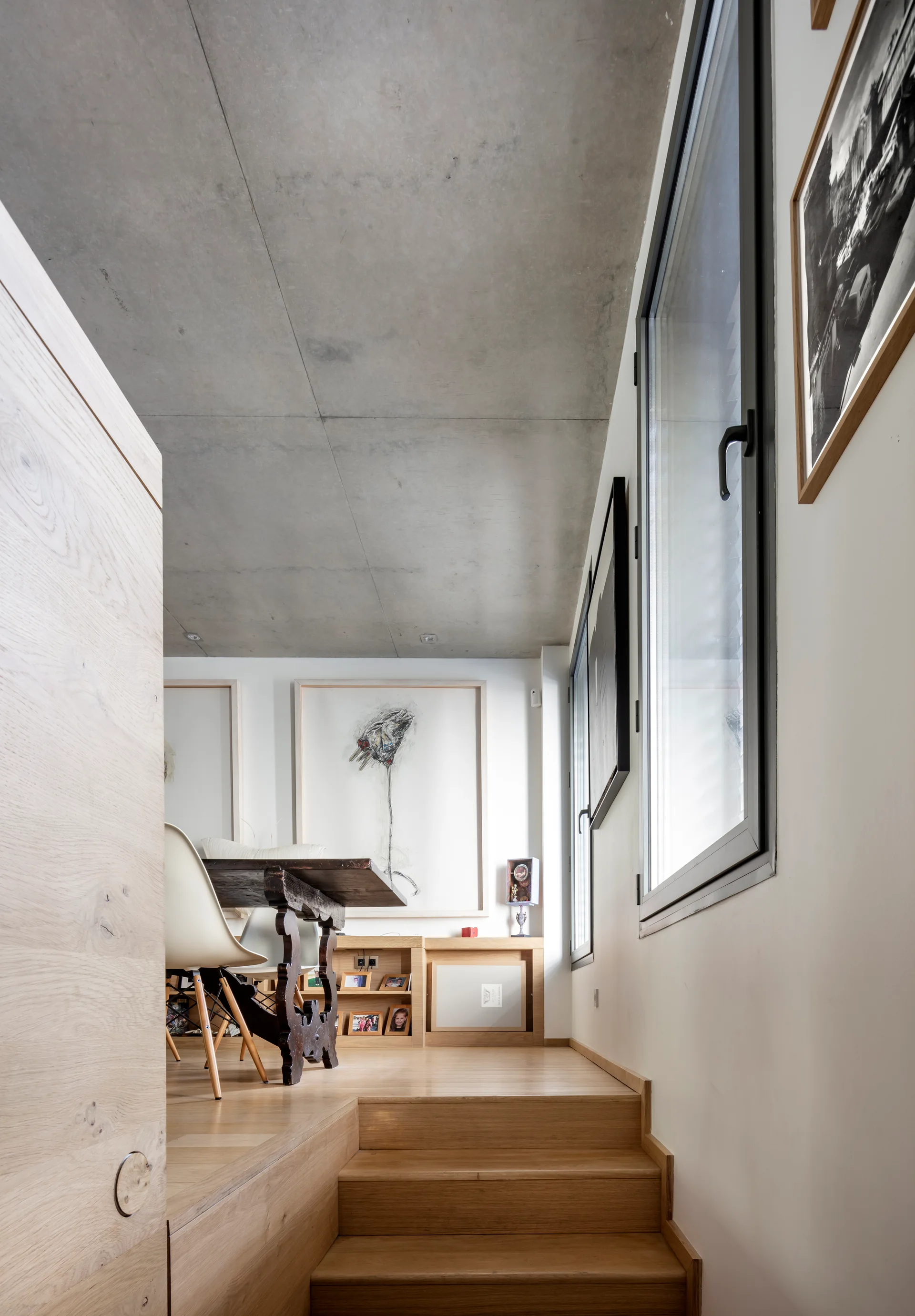 A split-level dining area features light wood stairs leading to a space with a dark wood table, white chairs, and an exposed concrete ceiling.