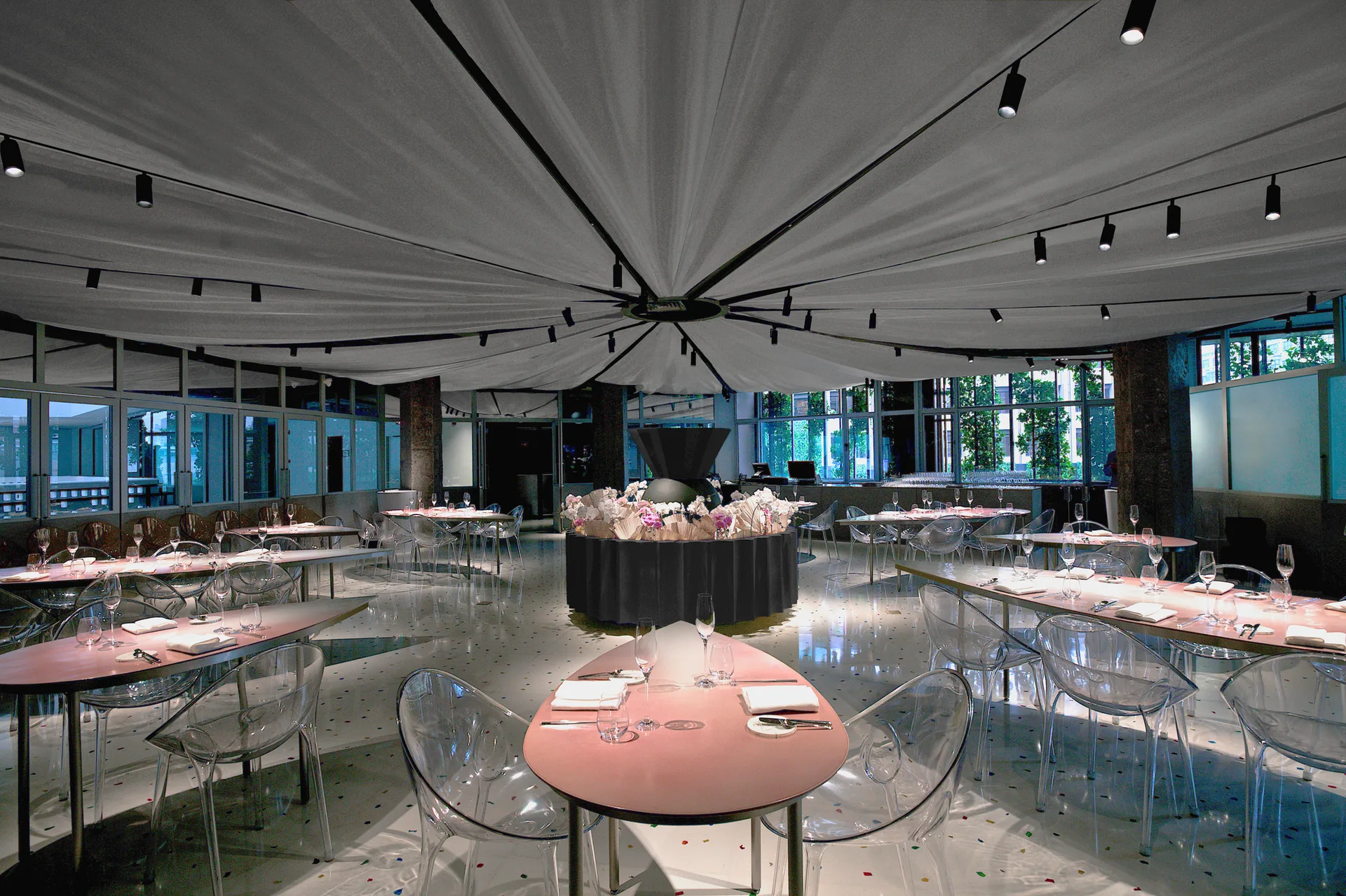 A spacious dining hall features teardrop-shaped tables, transparent plastic chairs, and a pleated white fabric ceiling canopy over a terrazzo floor.