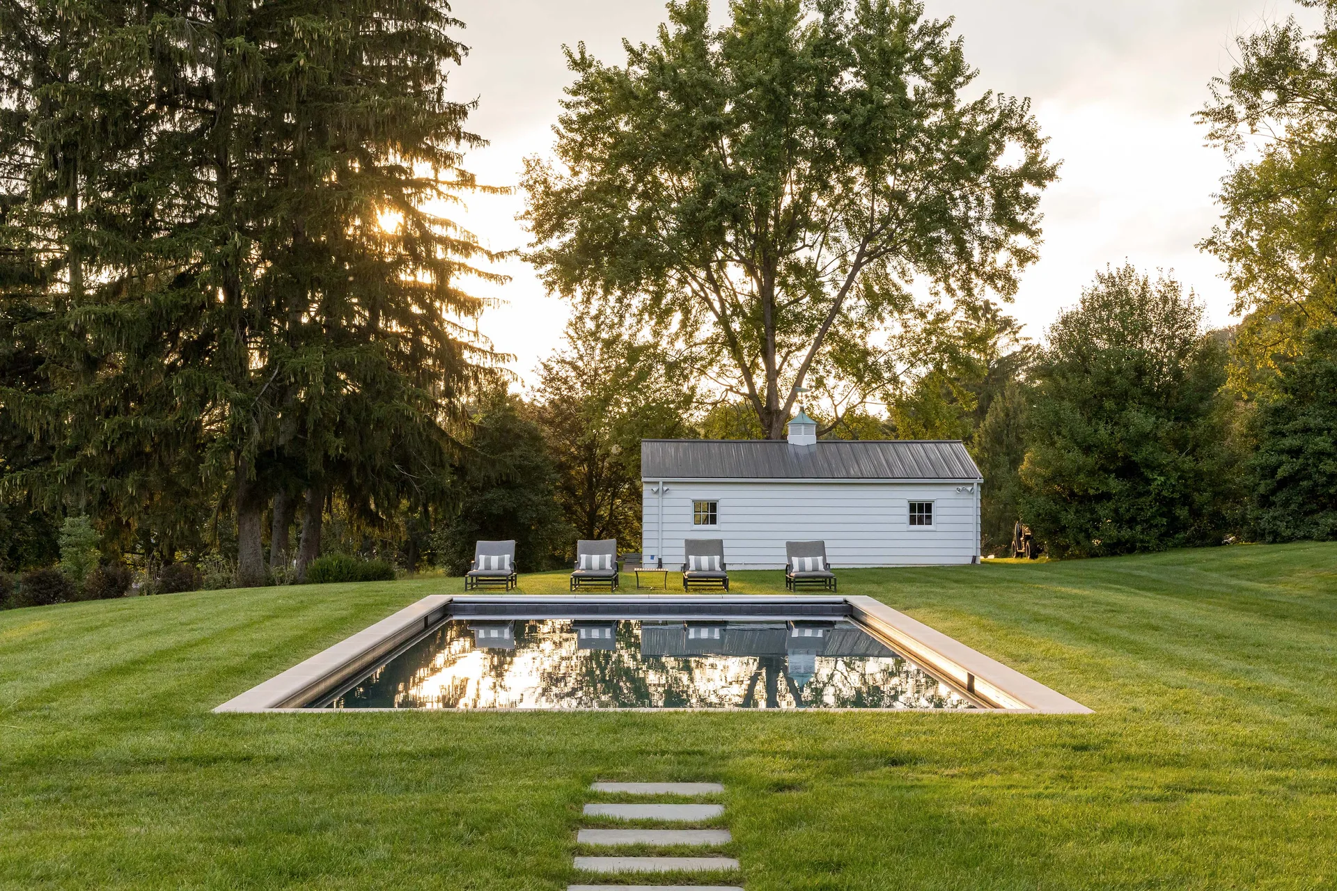 A rectangular swimming pool set directly in a green grass lawn with four lounge chairs and a white pool house in the background.