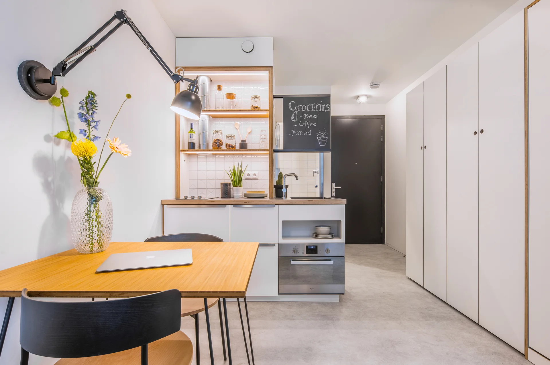 A minimalist studio kitchen features a compact white kitchenette, built-in open shelves with lighting, a wooden dining table, and tall white closets.