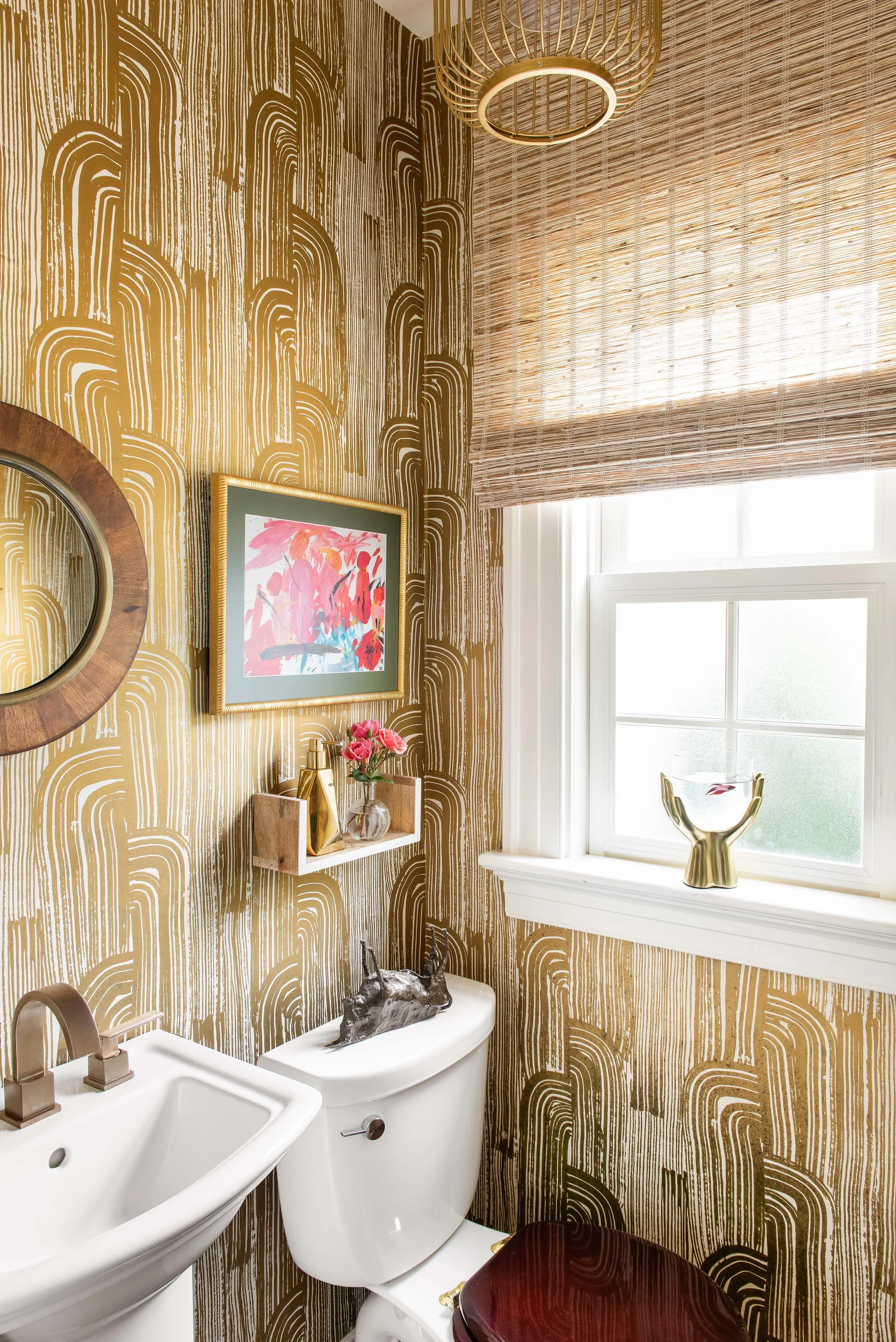 A small powder room features gold and white arch-patterned wallpaper, a white pedestal sink, and a woven bamboo window shade.