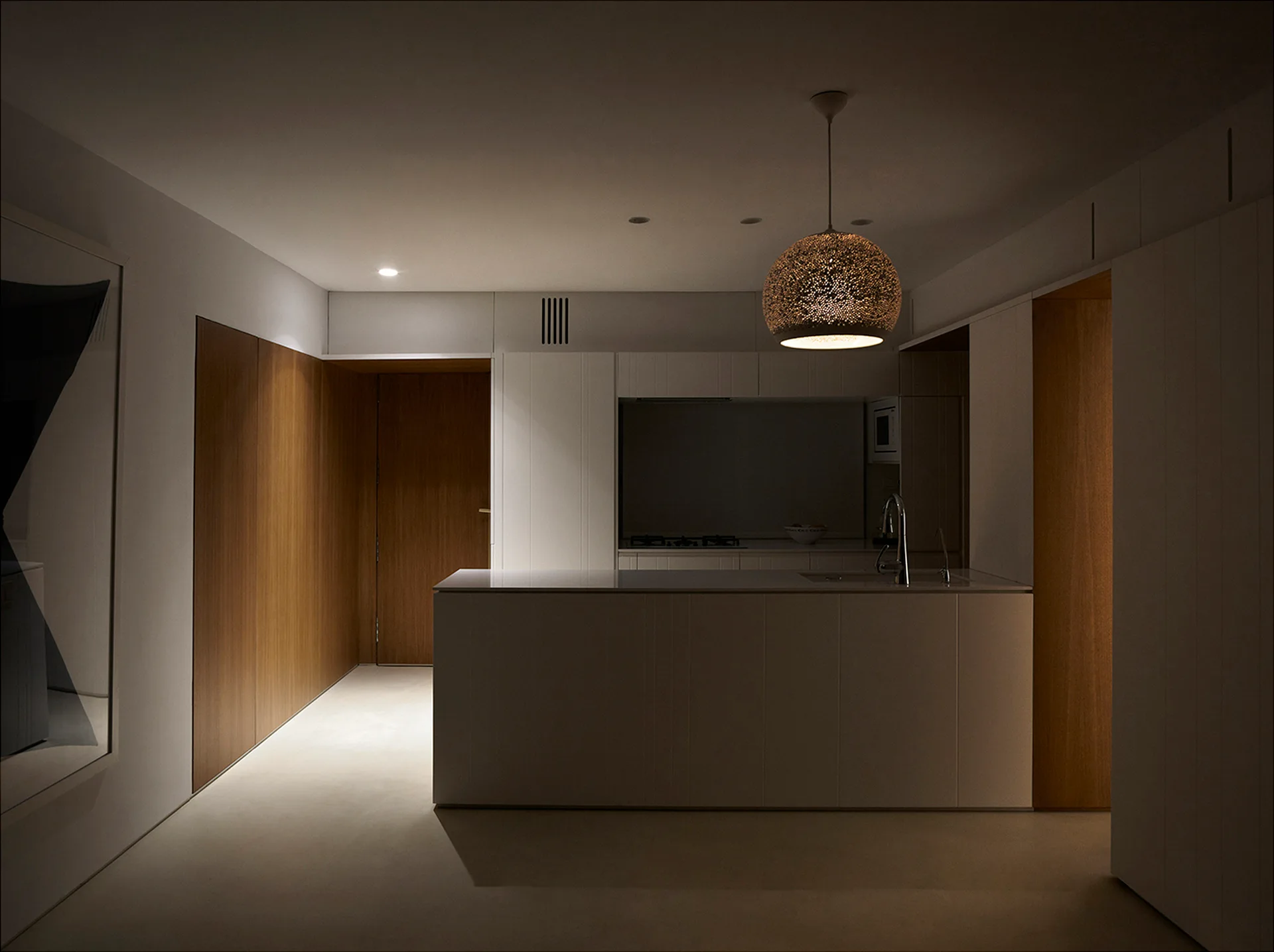 A minimalist kitchen features a white central island, flush white cabinetry, warm wood wall paneling, and a single dome pendant light casting a warm glow.