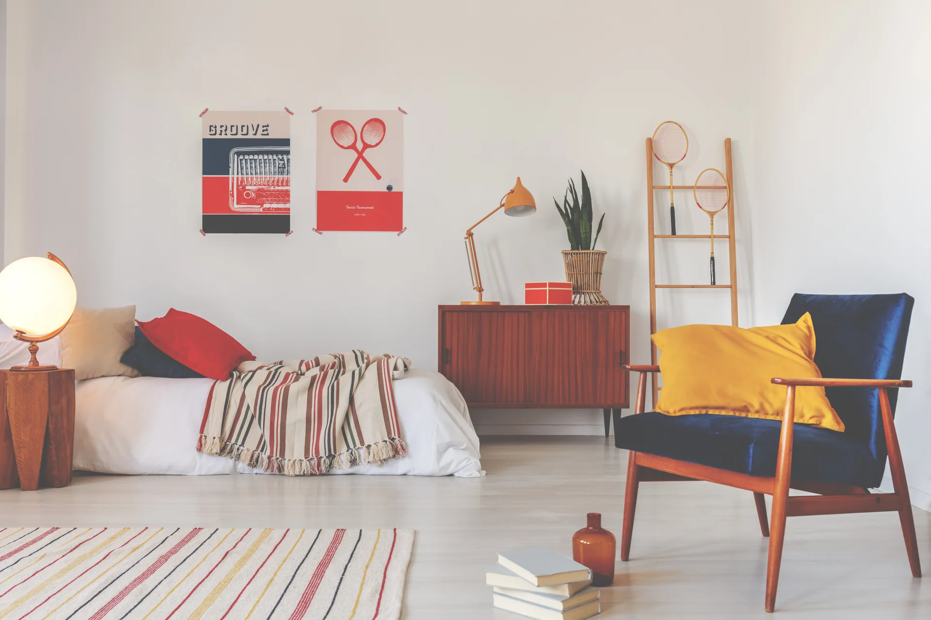 A low bed with a striped throw sits next to a wooden sideboard and a blue armchair with a yellow pillow in a bright room.
