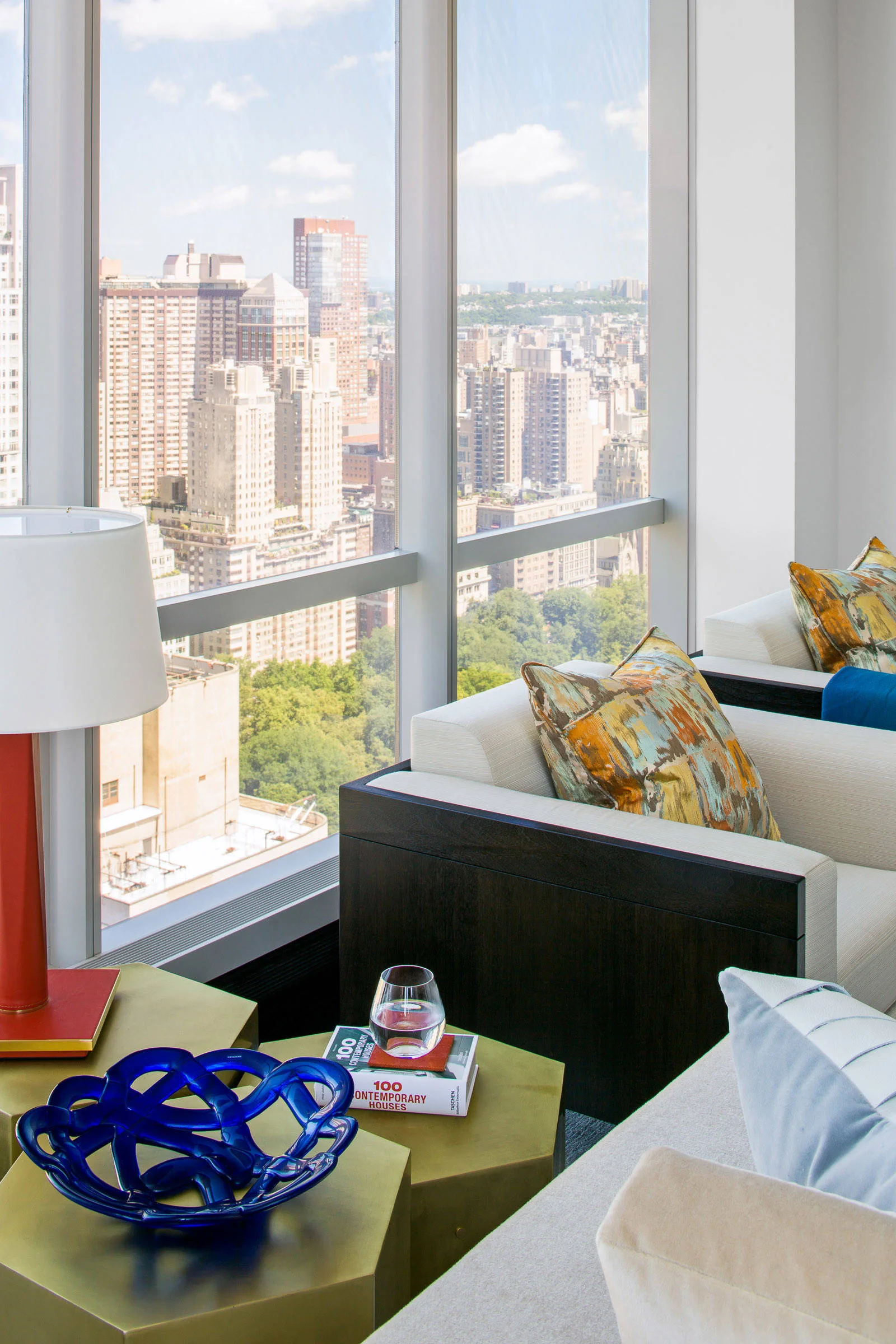 A contemporary high-rise living area with cream armchairs, a hexagonal brass coffee table, and a large window showing a cityscape view.