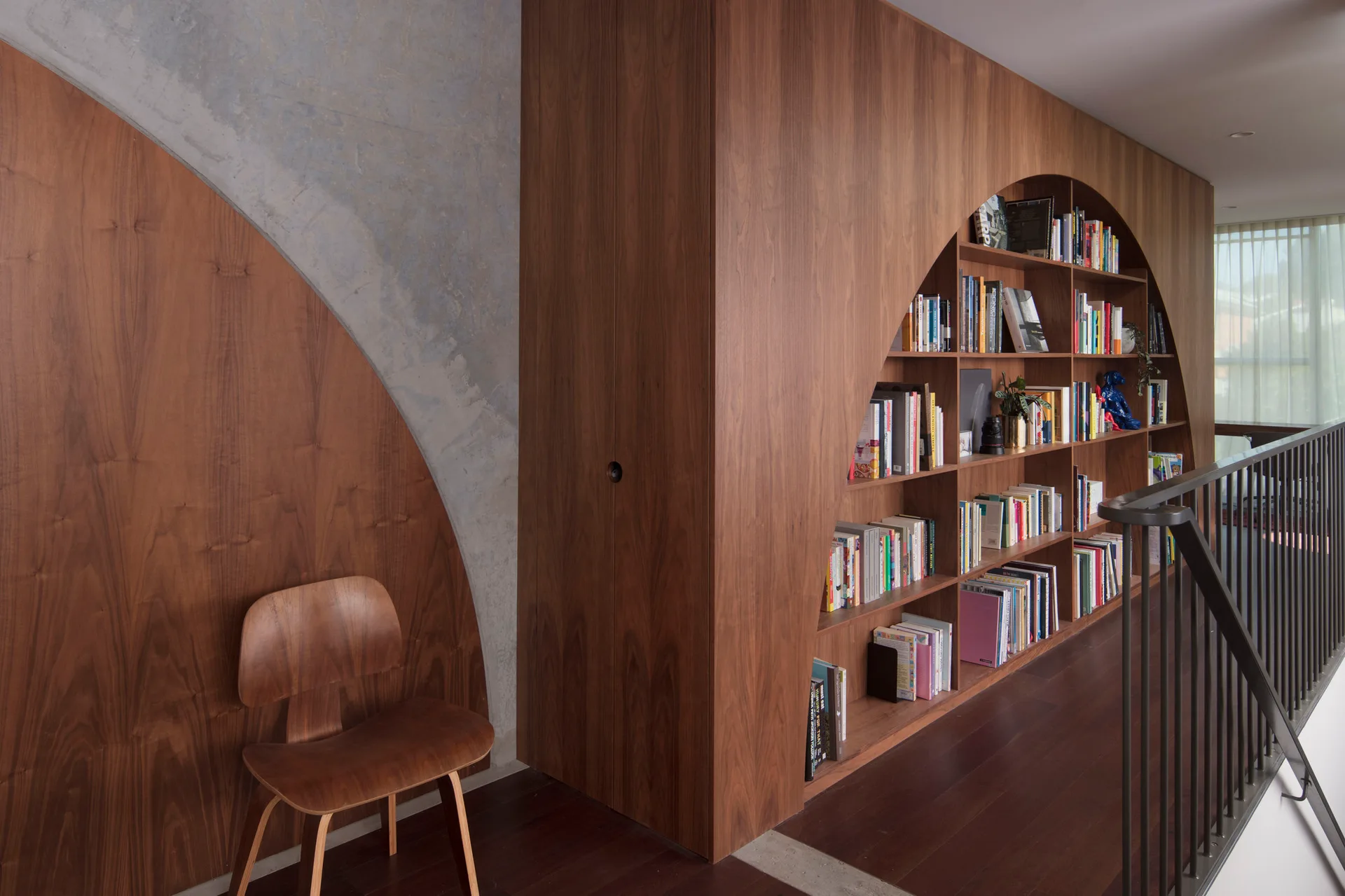 A custom curved wood bookshelf built into a hallway wall next to a matching plywood chair and metal stair railing on a landing.