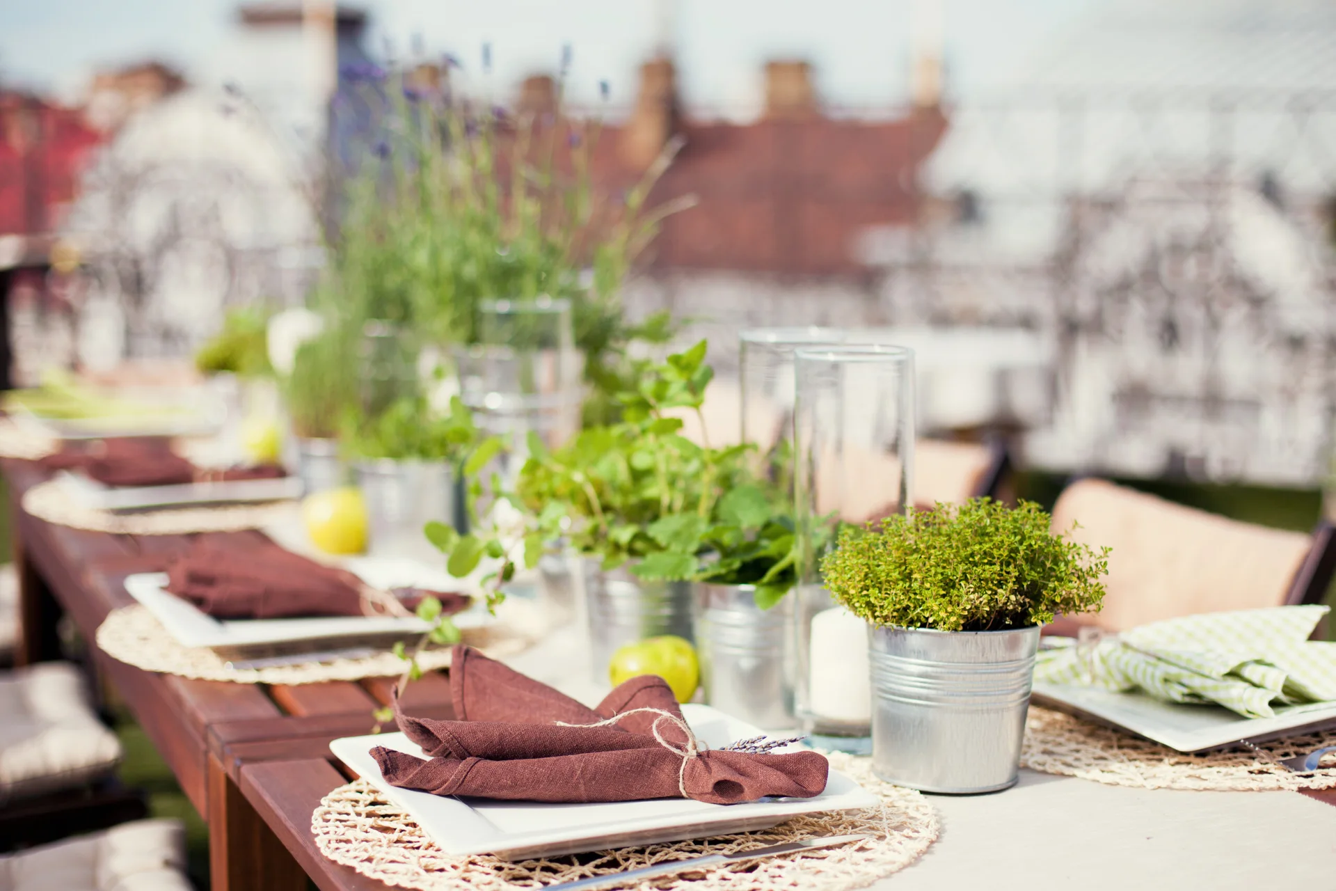 An outdoor wooden dining table is set with square white plates, woven circular placemats, brown napkins, and green herbs in galvanized metal pails.