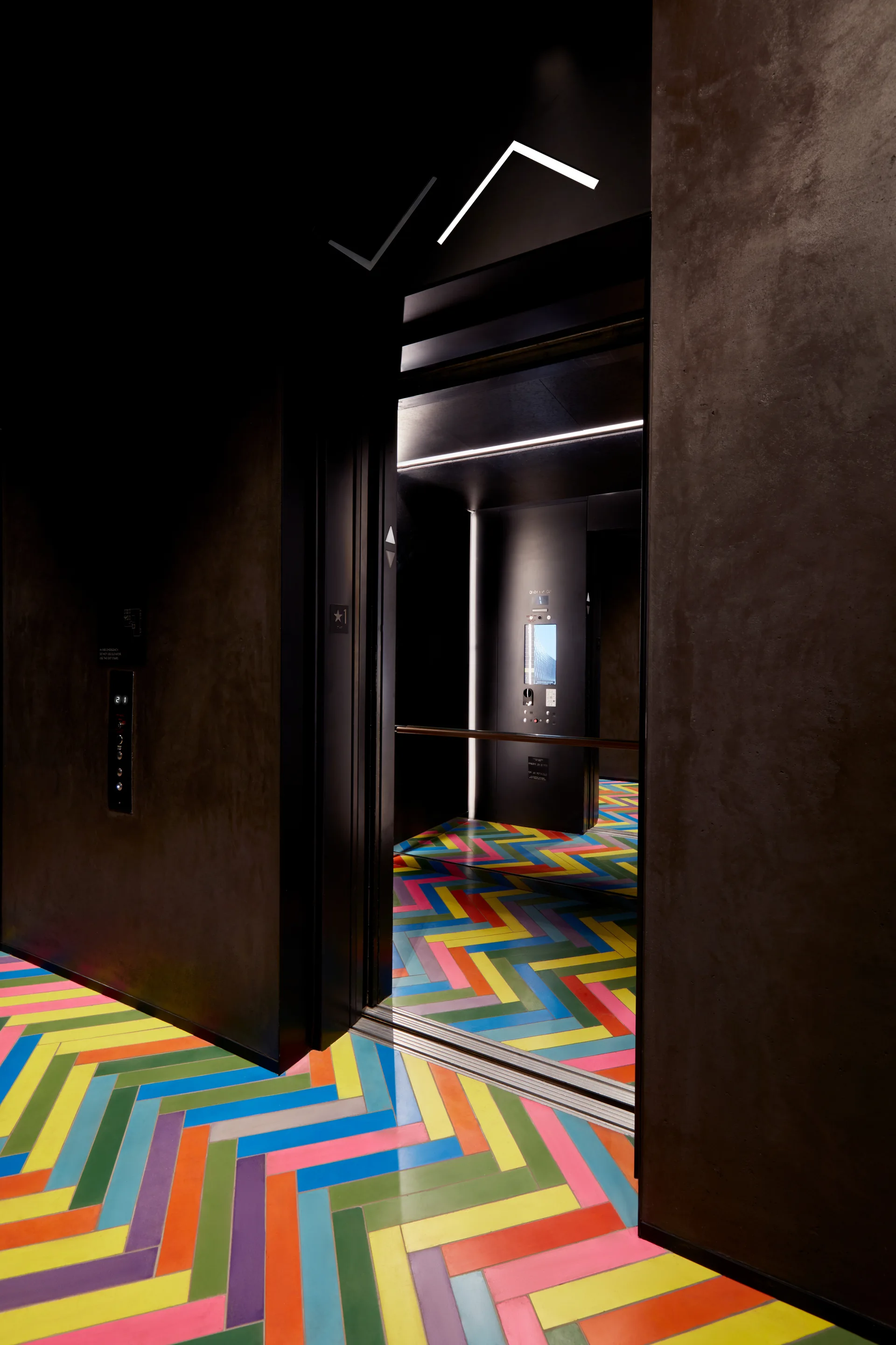 An elevator lobby with dark textured walls, an open elevator cab, and a bright multi-colored herringbone patterned tile floor.
