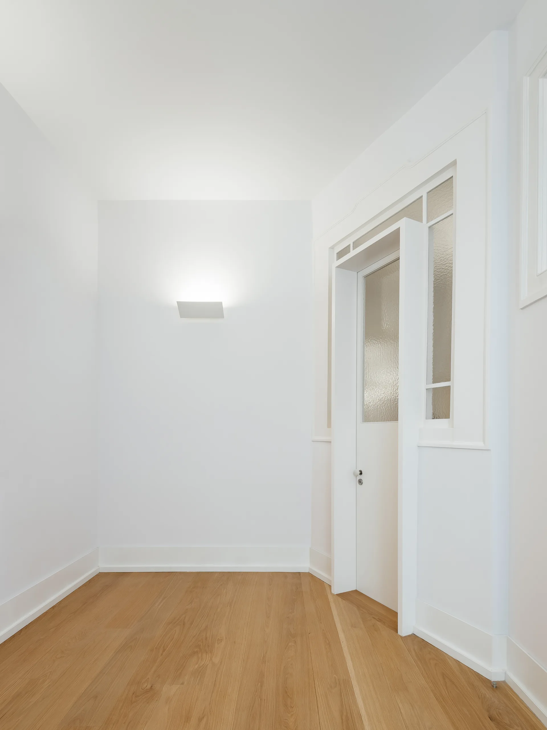 A minimalist white hallway features light oak floors, a white door with textured glass panels, and a modern rectangular wall sconce.