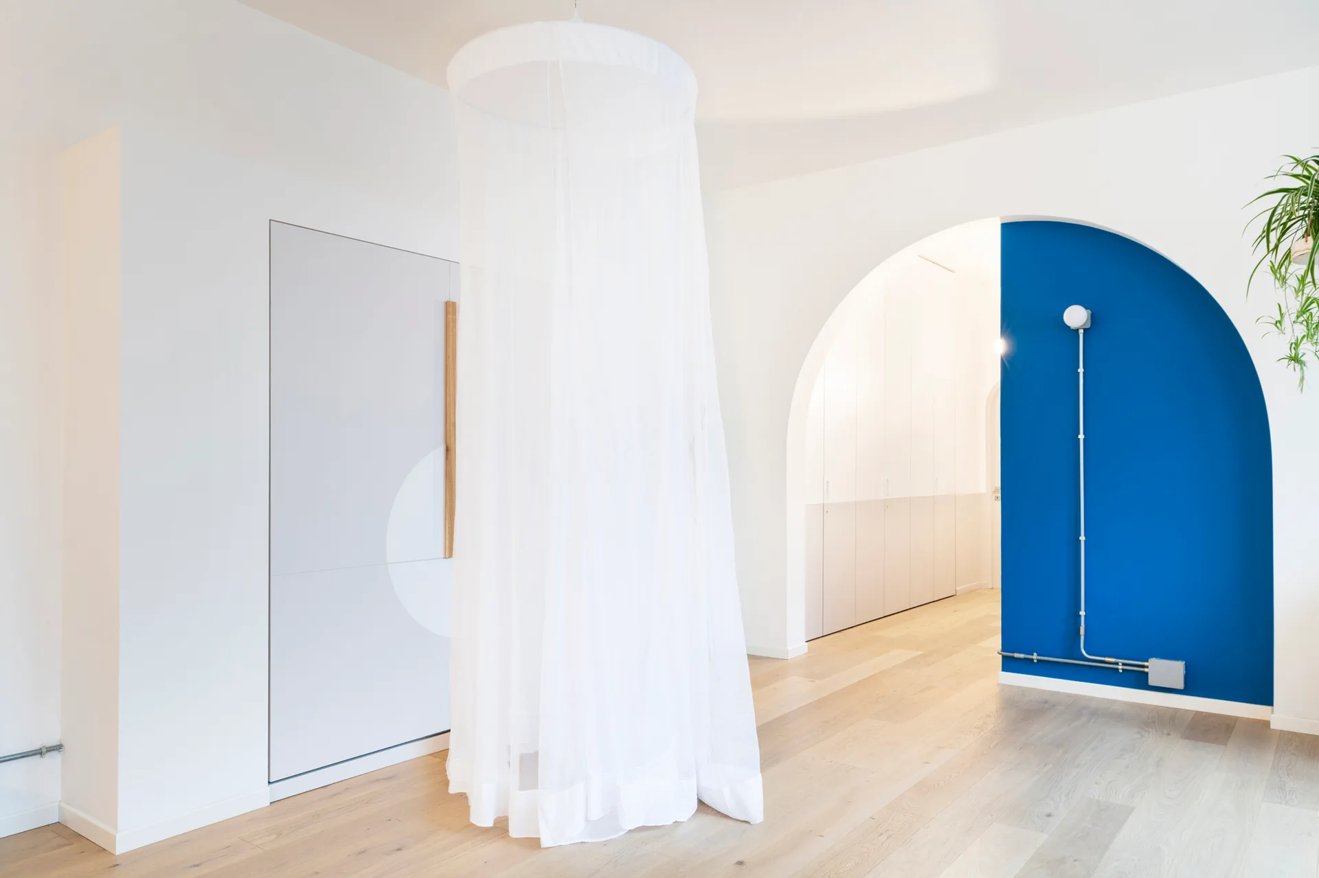 A minimalist white room features a hanging sheer canopy on light wood floors near a vivid blue arched wall niche and an open archway.