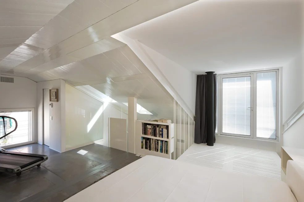 A bright minimalist attic bedroom with a white bed, slanted wood-planked ceilings, a grey floor, a built-in bookshelf, and a treadmill.