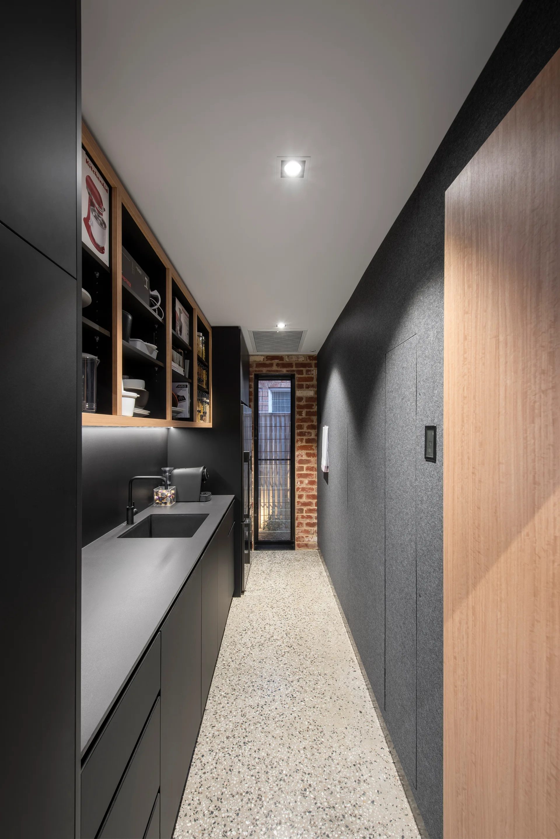 A narrow galley kitchen features matte black cabinetry, a speckled terrazzo floor, a textured grey wall, and a vertical window framed in exposed brick.