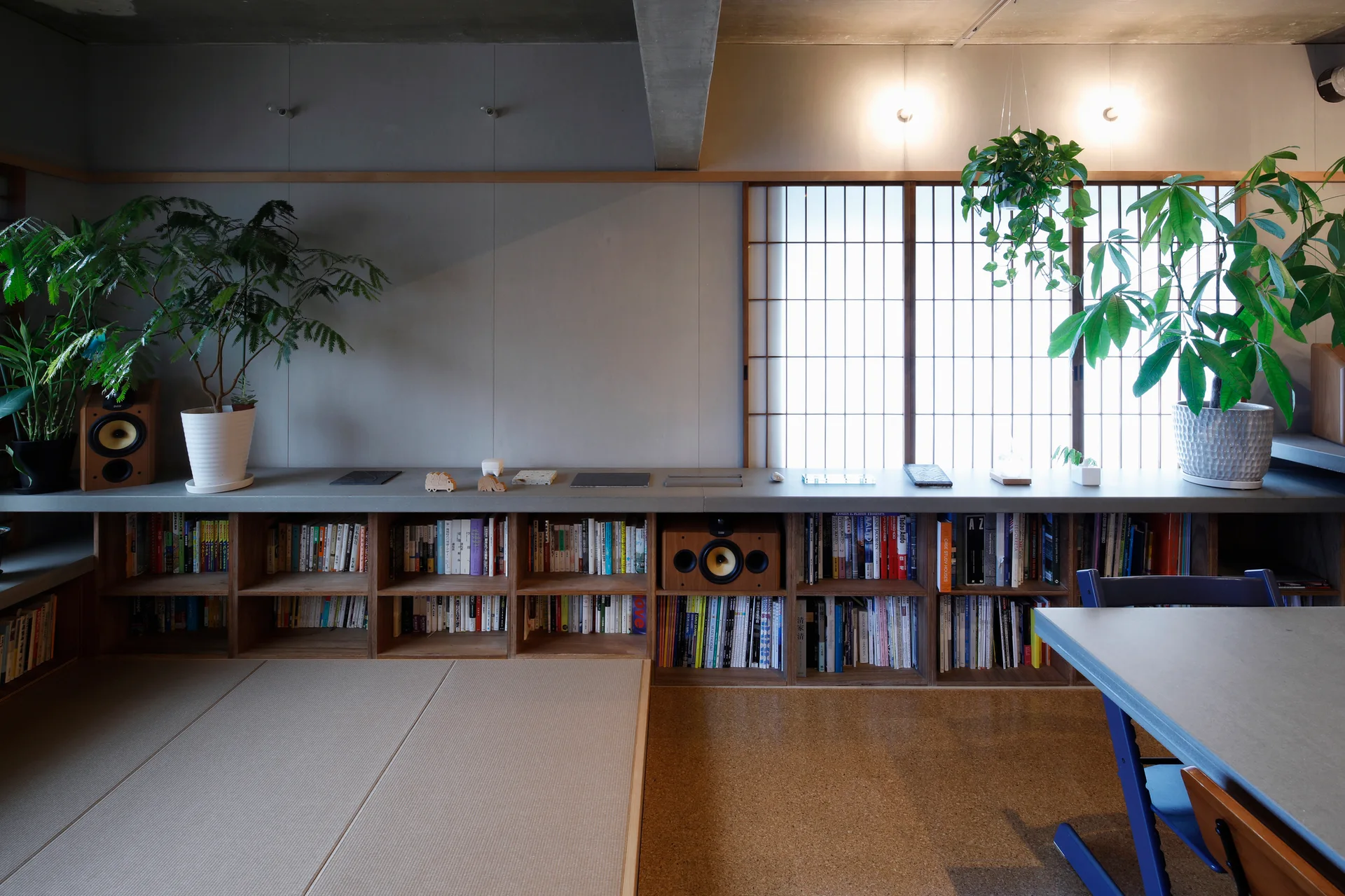 A minimalist room features a long low wooden bookshelf, a concrete countertop, tatami mats, potted plants, and a shoji screen window.