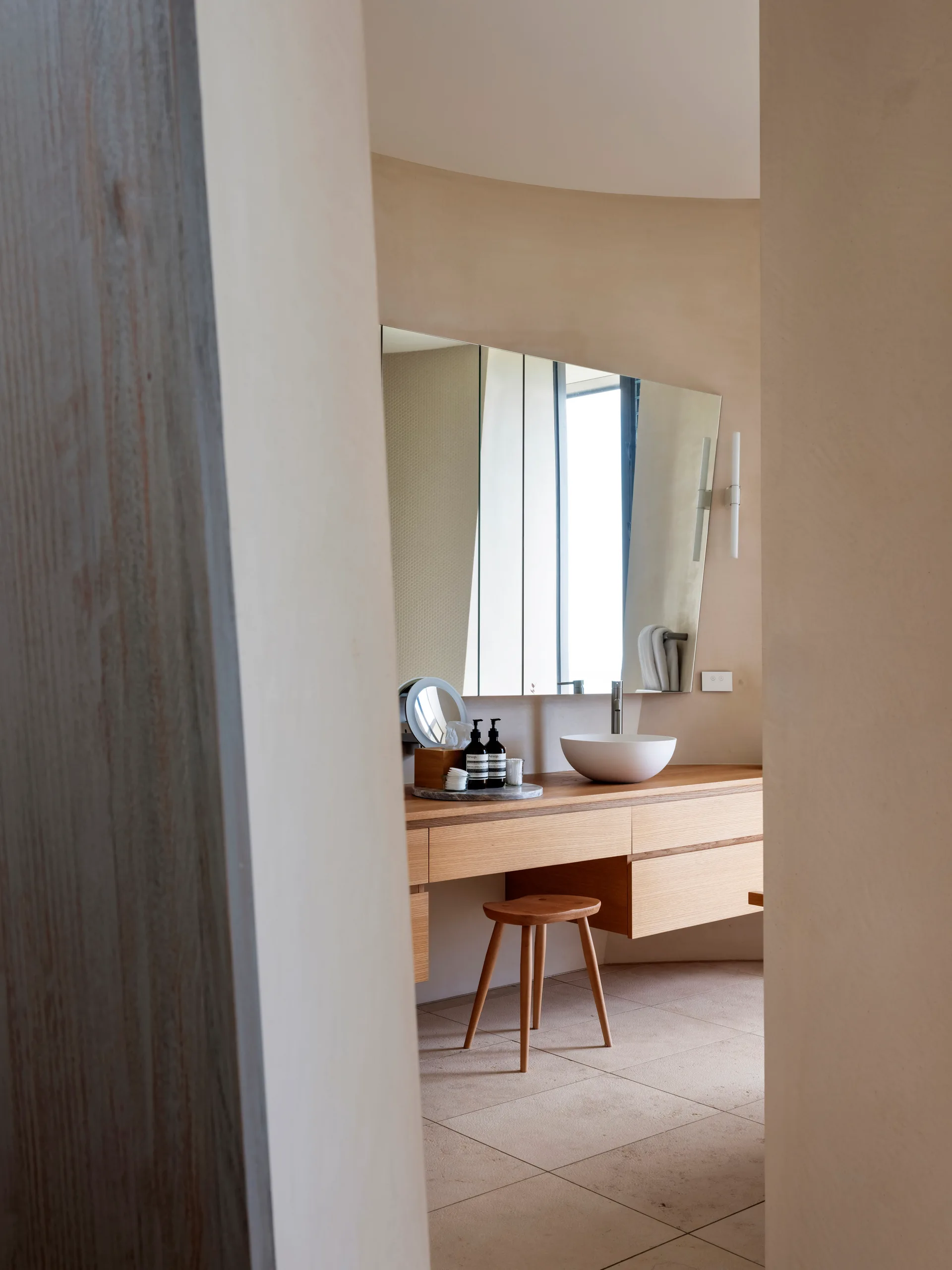 A floating wooden bathroom vanity with a white vessel sink and matching stool set against curved beige plaster walls.