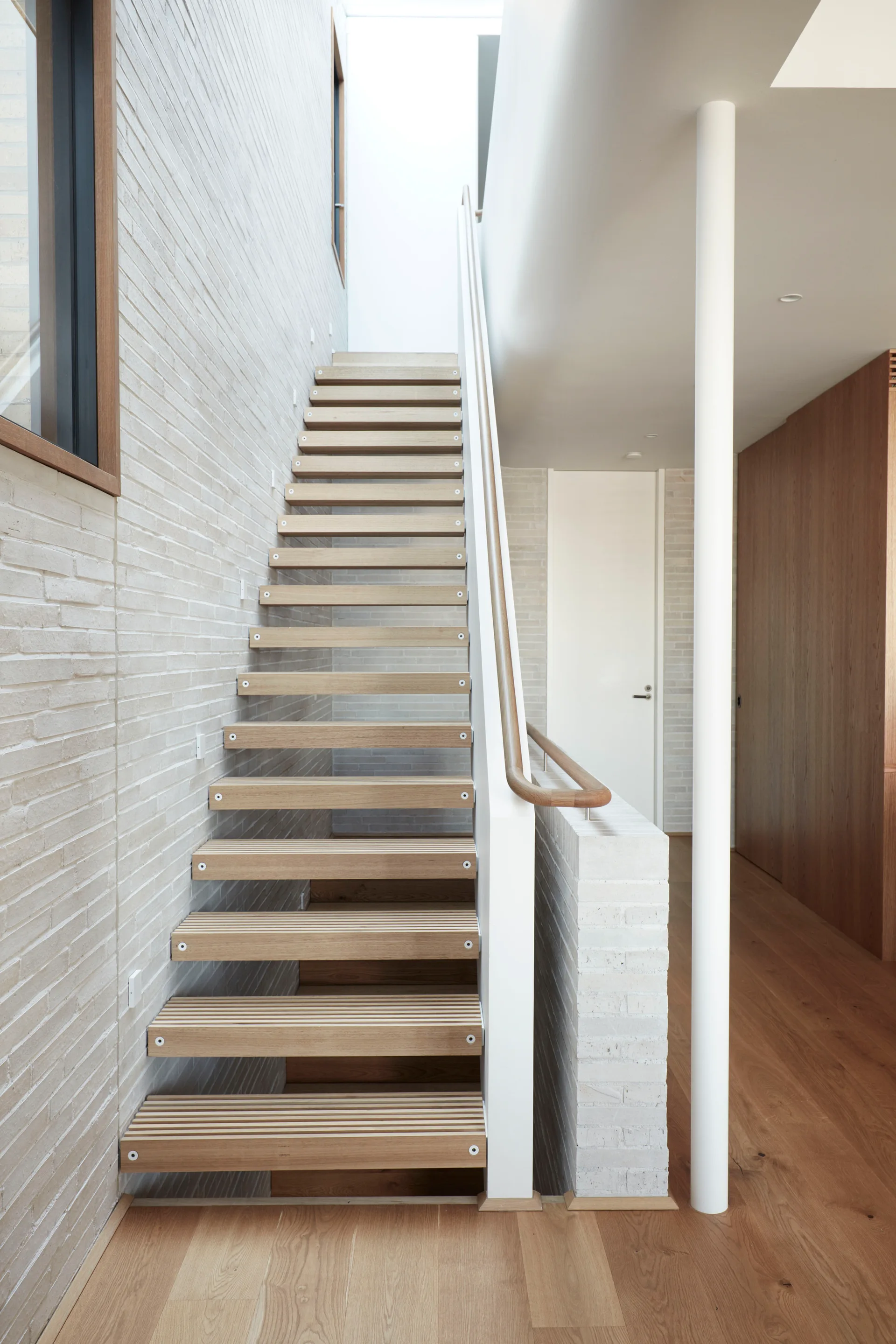 A modern minimalist staircase features floating wood treads, a matching wooden handrail, and a textured light grey brick wall.