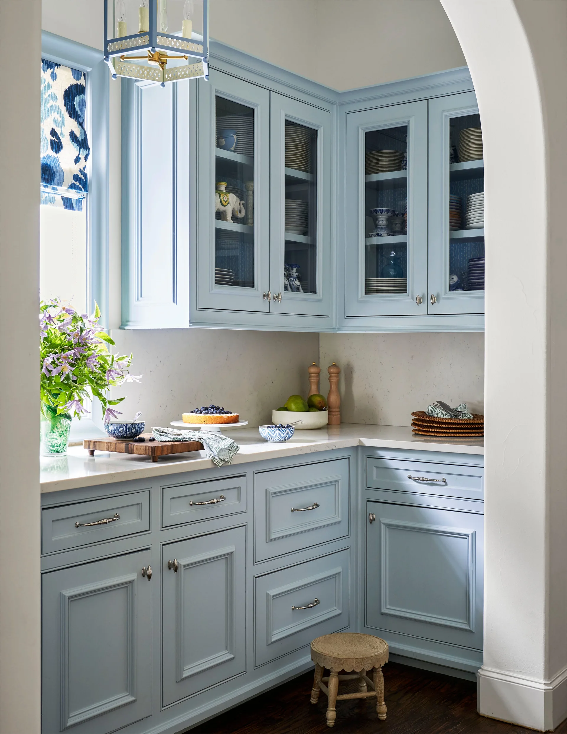 A pale blue butler's pantry with glass-front upper cabinets, white stone countertops, and a wooden stool, viewed through a white plastered archway.