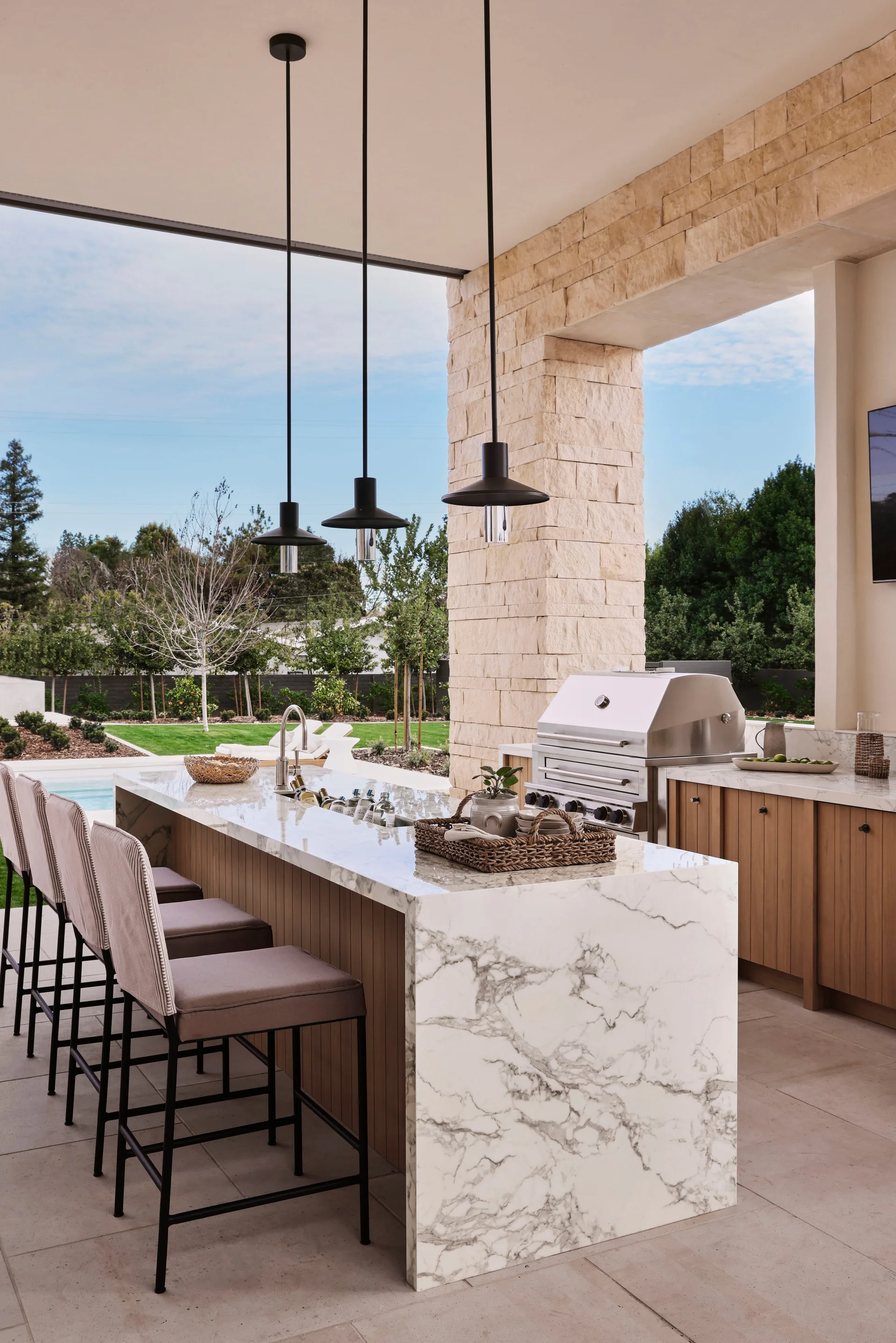 An outdoor kitchen features a white marble waterfall island with four bar stools beneath black pendant lights next to a built-in stainless steel grill.