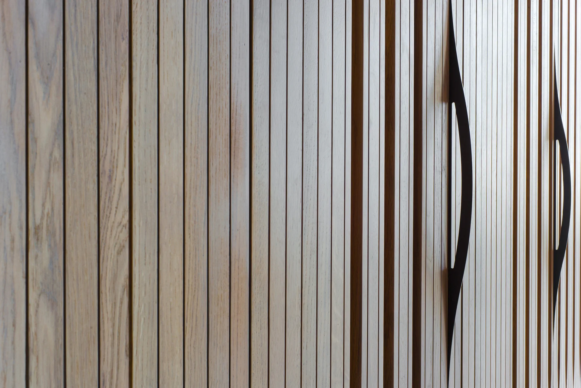 A close-up of light brown slatted wood cabinet doors featuring prominent, curved black metal pull handles integrated into the vertical grooves.