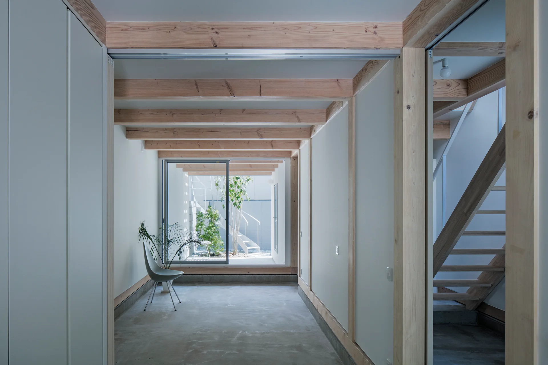 A minimalist hallway features exposed light-wood ceiling beams, concrete flooring, and a large sliding glass door revealing an enclosed courtyard.