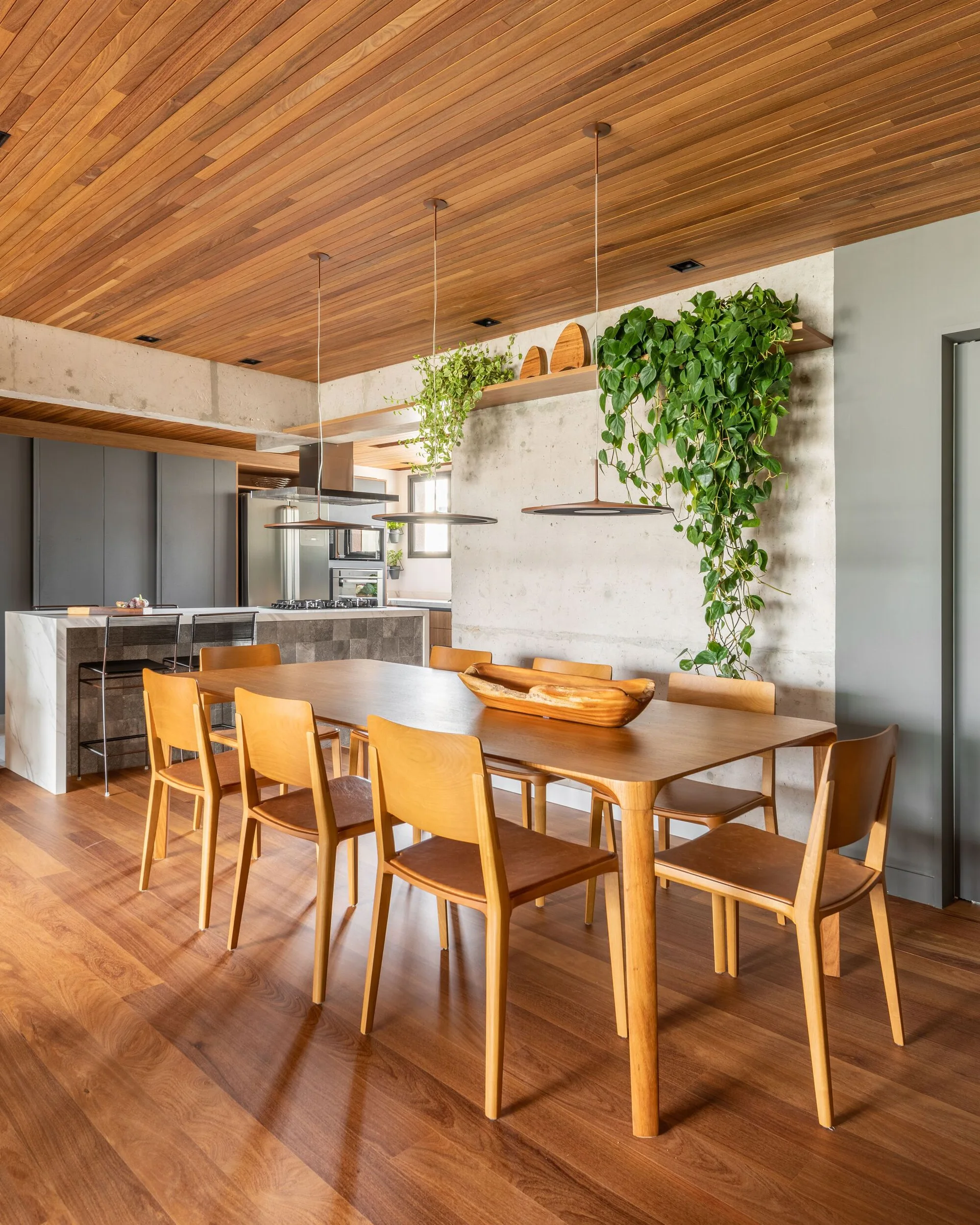A wooden dining set stands on wood flooring beneath a wood-slat ceiling, opposite a raw concrete wall decorated with a trailing green plant.