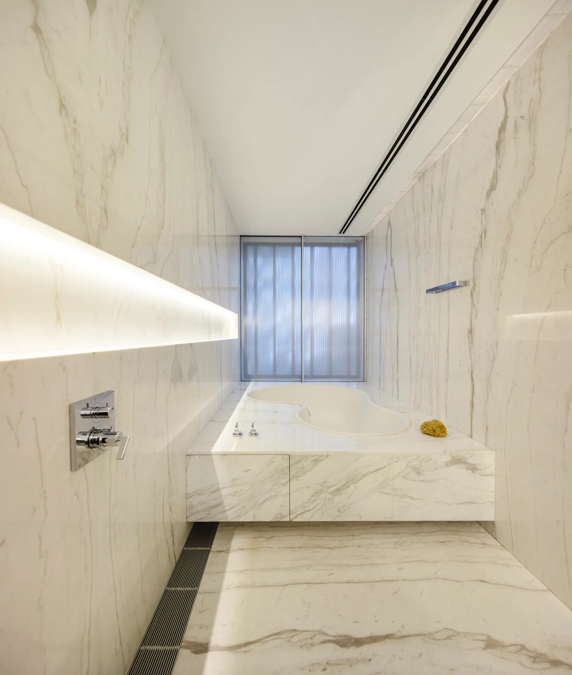 A minimalist narrow bathroom fully clad in white veined marble featuring a curvilinear built-in bathtub and a soft linear cove light.