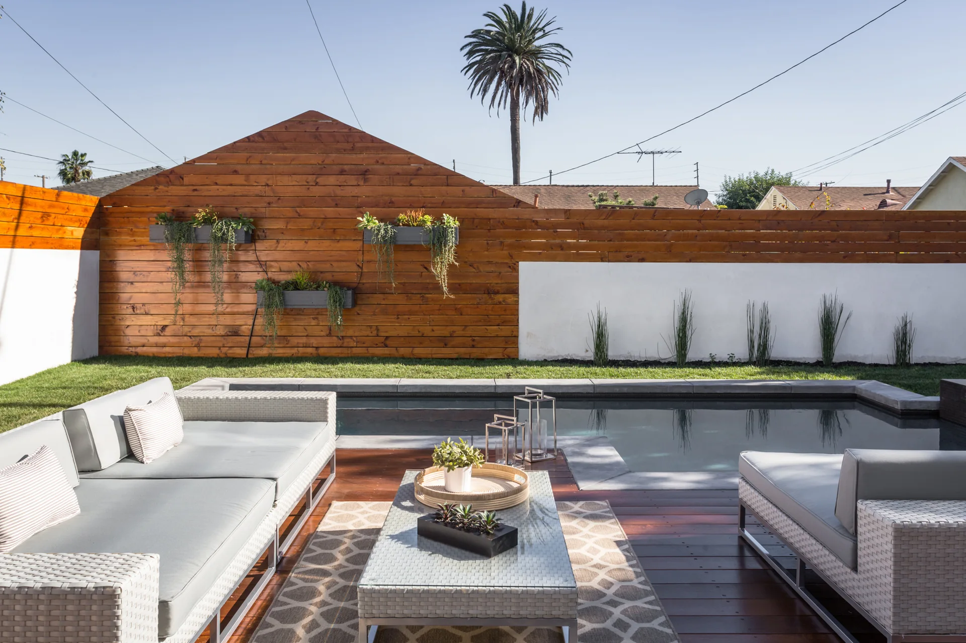 A modern backyard features grey wicker patio furniture on a wood deck next to a swimming pool and a wood and white stucco fence.