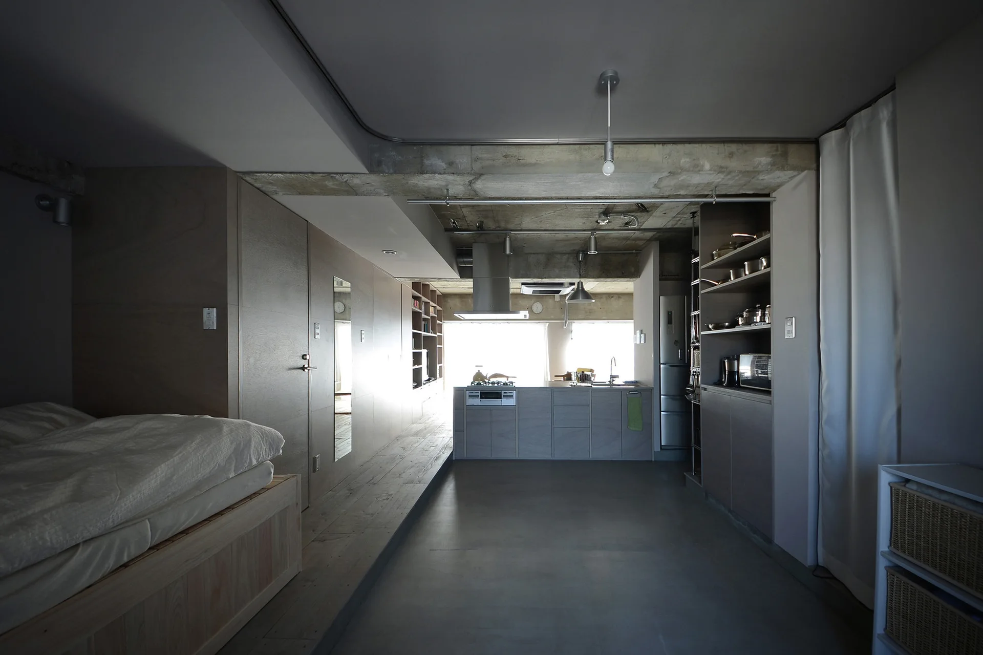 A modern industrial studio apartment featuring a low wooden platform bed, a minimalist kitchen island, and an exposed concrete ceiling.