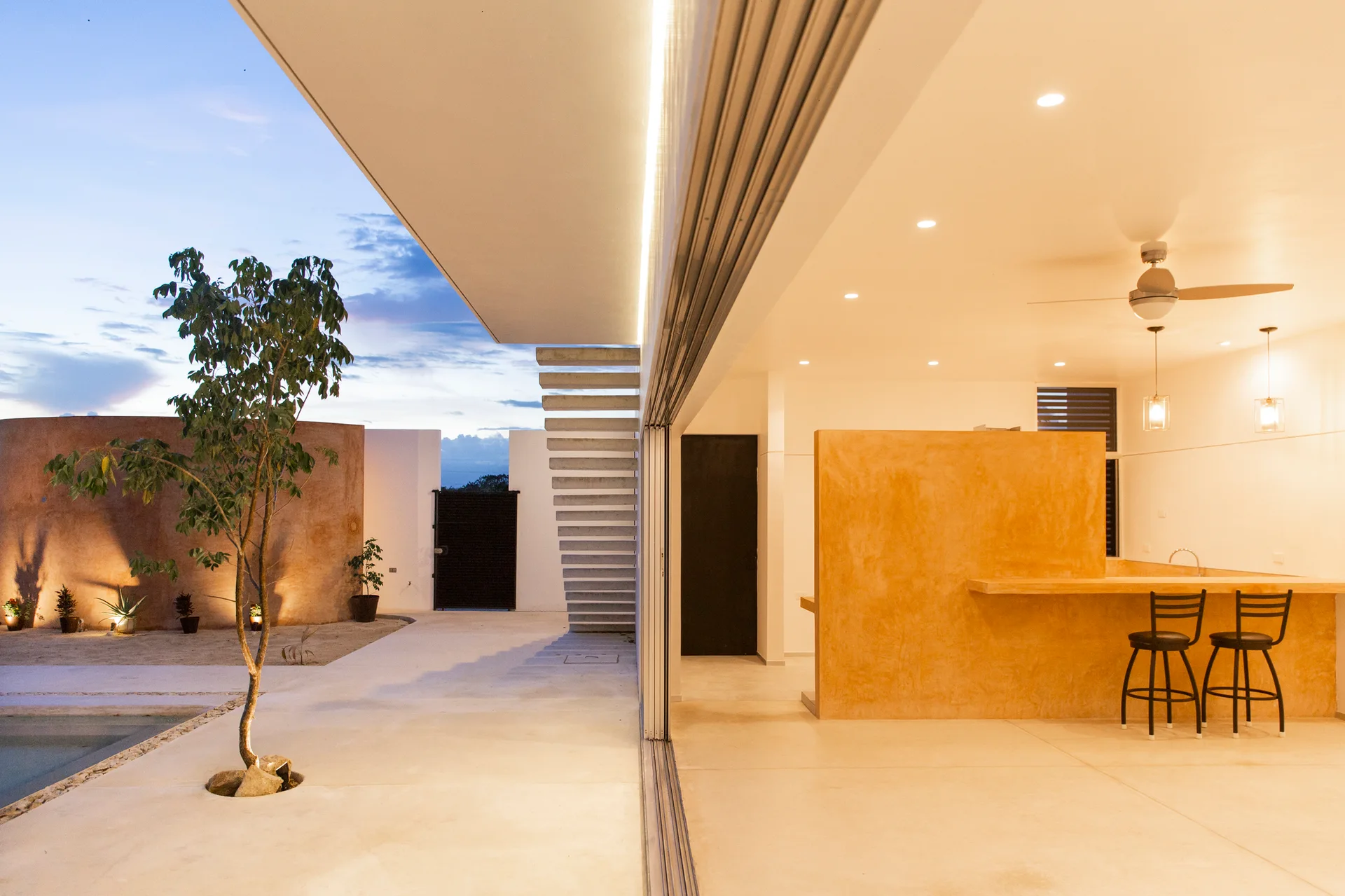 A modern indoor-outdoor kitchen featuring an ochre plaster island with bar stools, opening onto a concrete patio with floating stairs and a tree.