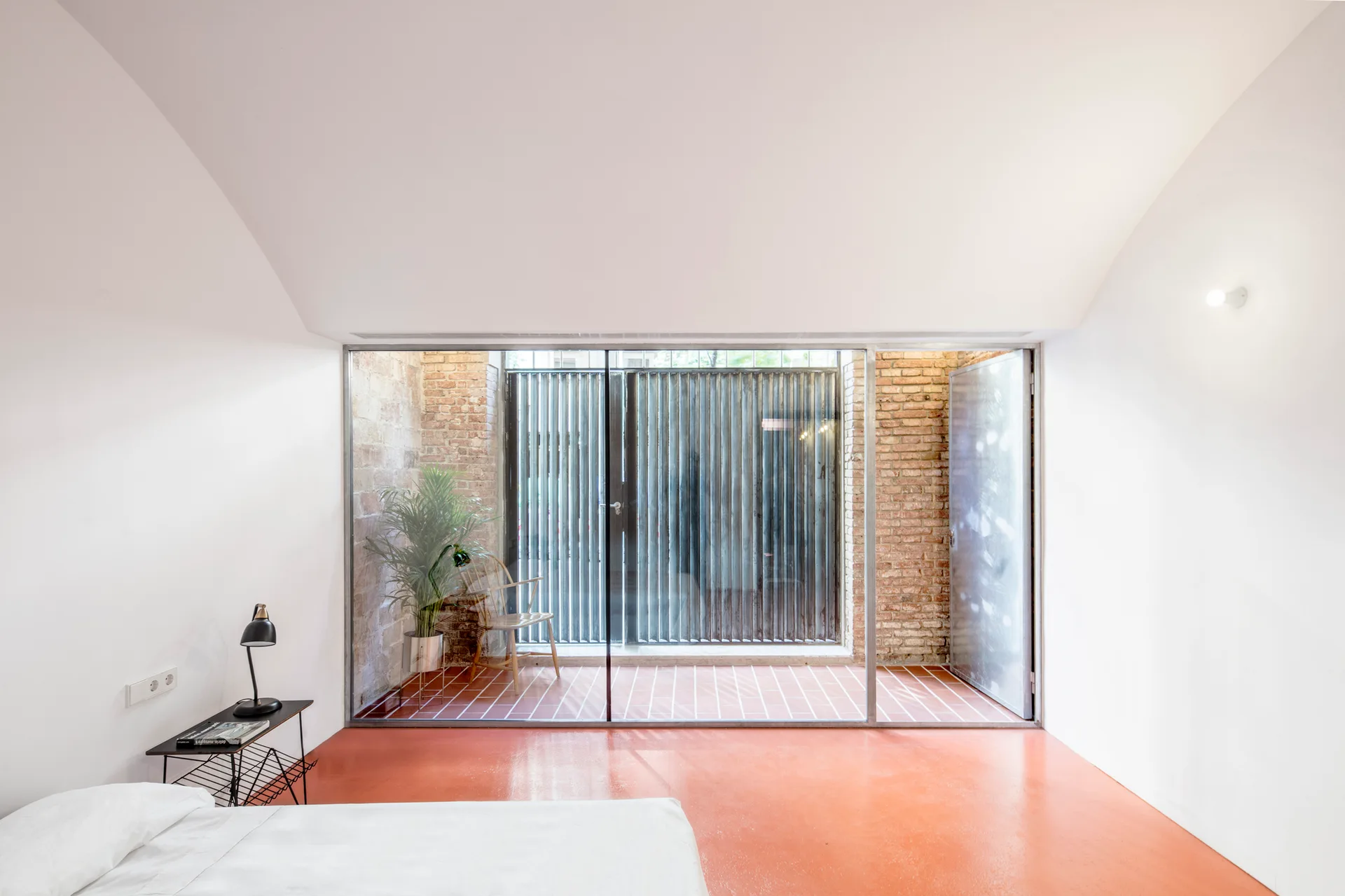 A minimalist bedroom featuring a vibrant orange floor and a large glass partition looking out onto a brick patio with a single chair and plant.