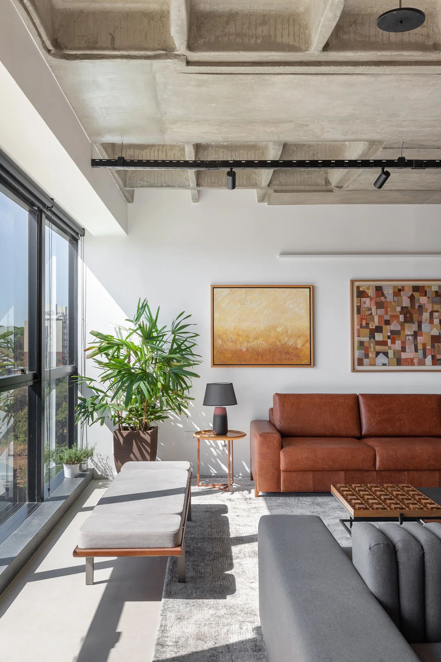 A modern living room features a brown leather sofa, a gray upholstered bench, a large green houseplant, and an exposed concrete waffle ceiling.