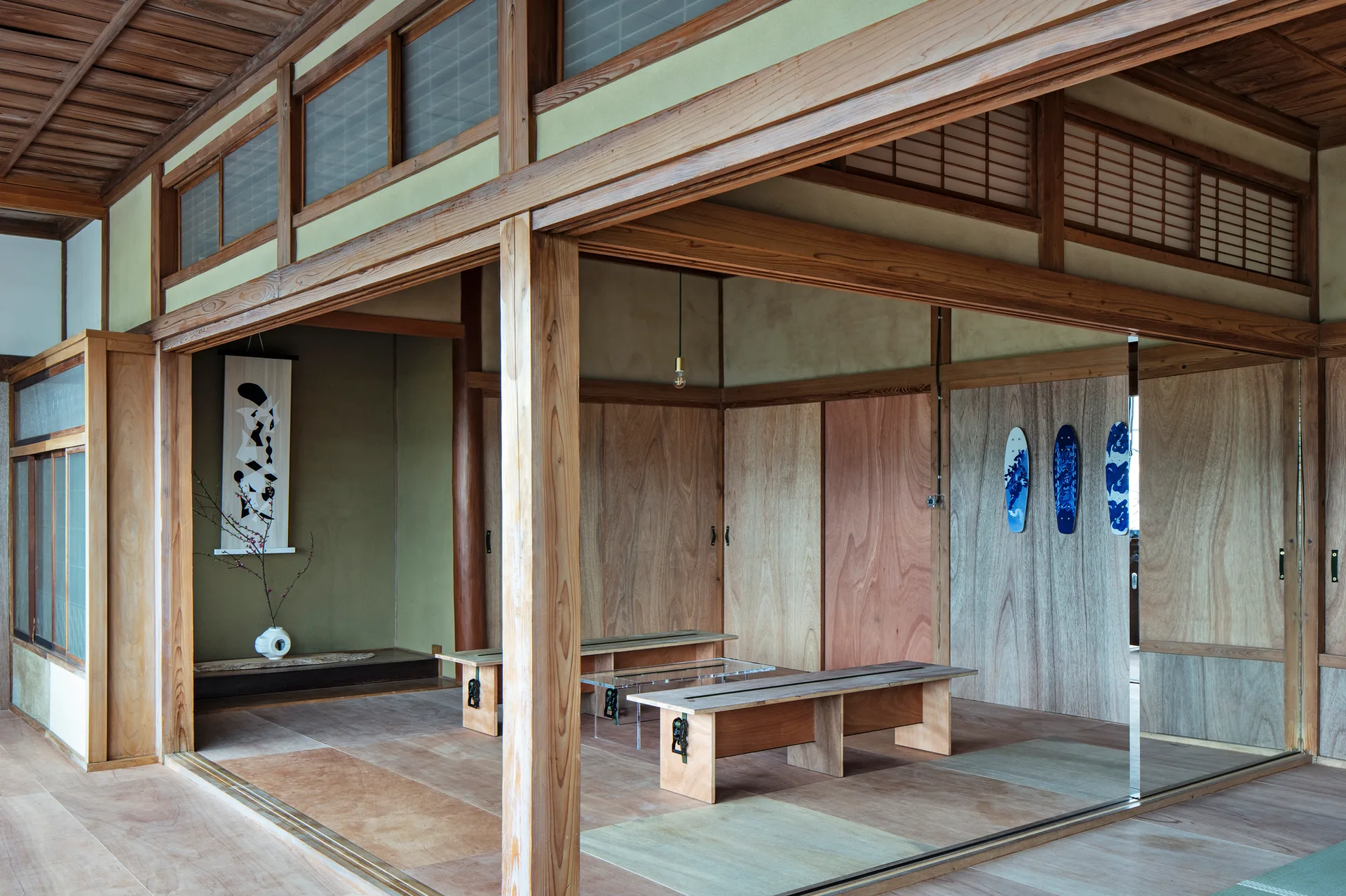 A Japanese-style room features exposed timber beams, light plywood wall paneling, two wooden benches, and three blue skateboards hanging on the wall.