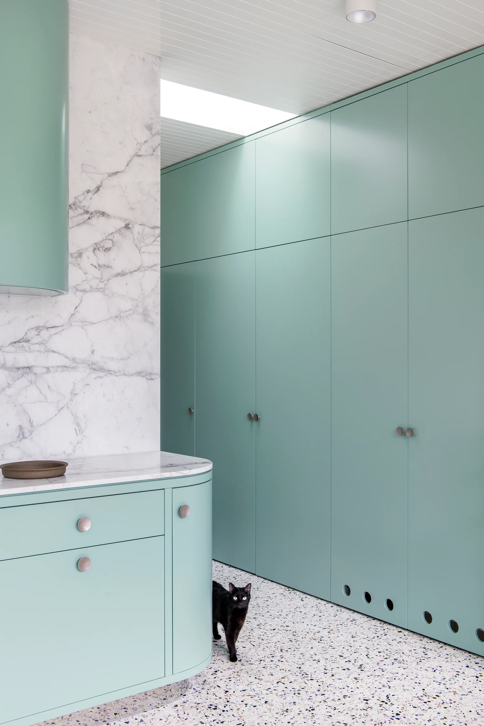 A minimalist kitchen space features full-height mint green cabinets, a white veined marble wall, and multi-colored speckled terrazzo flooring.